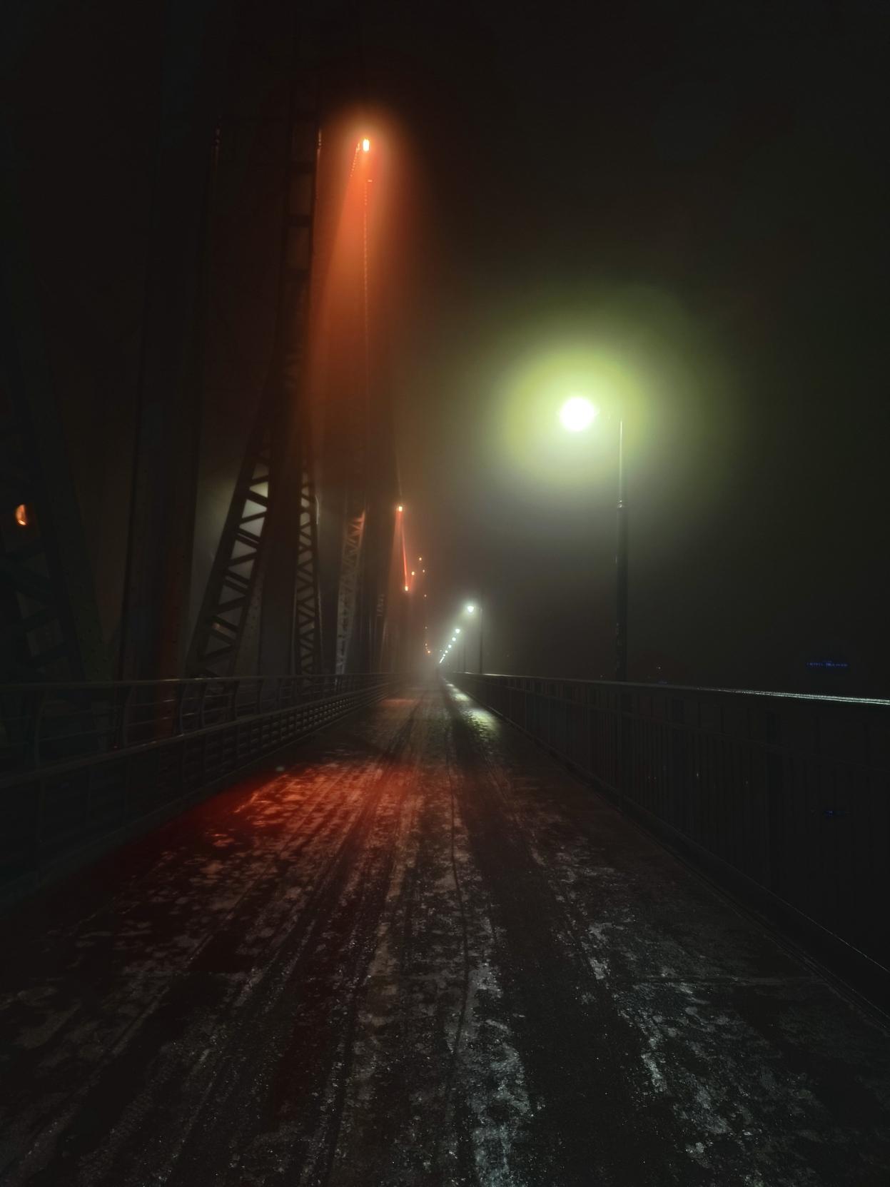 Zaśnieżony chodnik na moście Józefa Piłsudskiego. We mgle widoczne oświetlone na czerwono przęsła mostu oraz latarnie przy barierce.