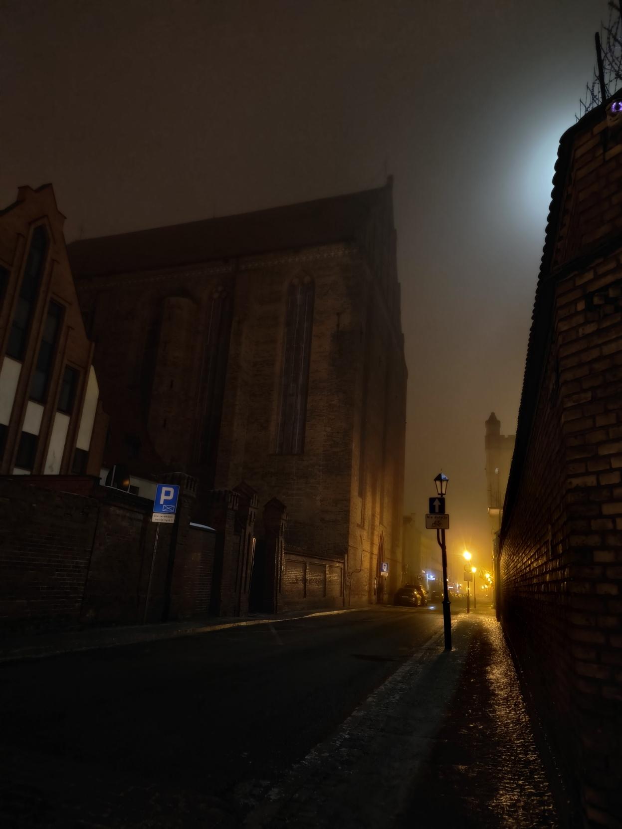 Kościół Mariacki w Toruniu w nocnej mgle. Widoczna poświata księżyca oraz świateł ulicznych.