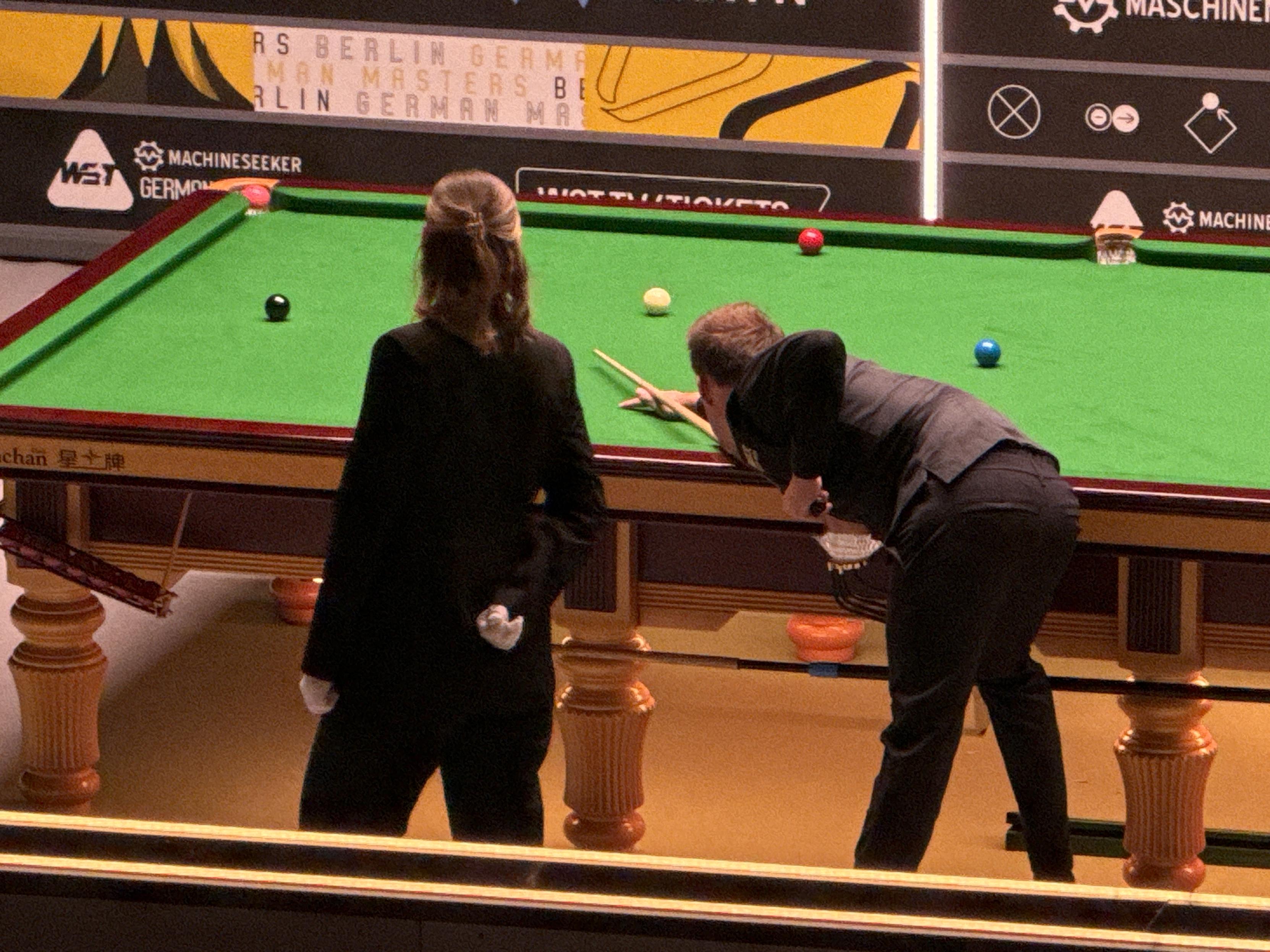 Mecz snookera, ćwierćfinał German Masters. Judd Trump, pochylony nad stołem, w trakcie wbicia różowej bili do lewej dolnej kieszeni. Za nim po lewej stoi sędzina spotkania.