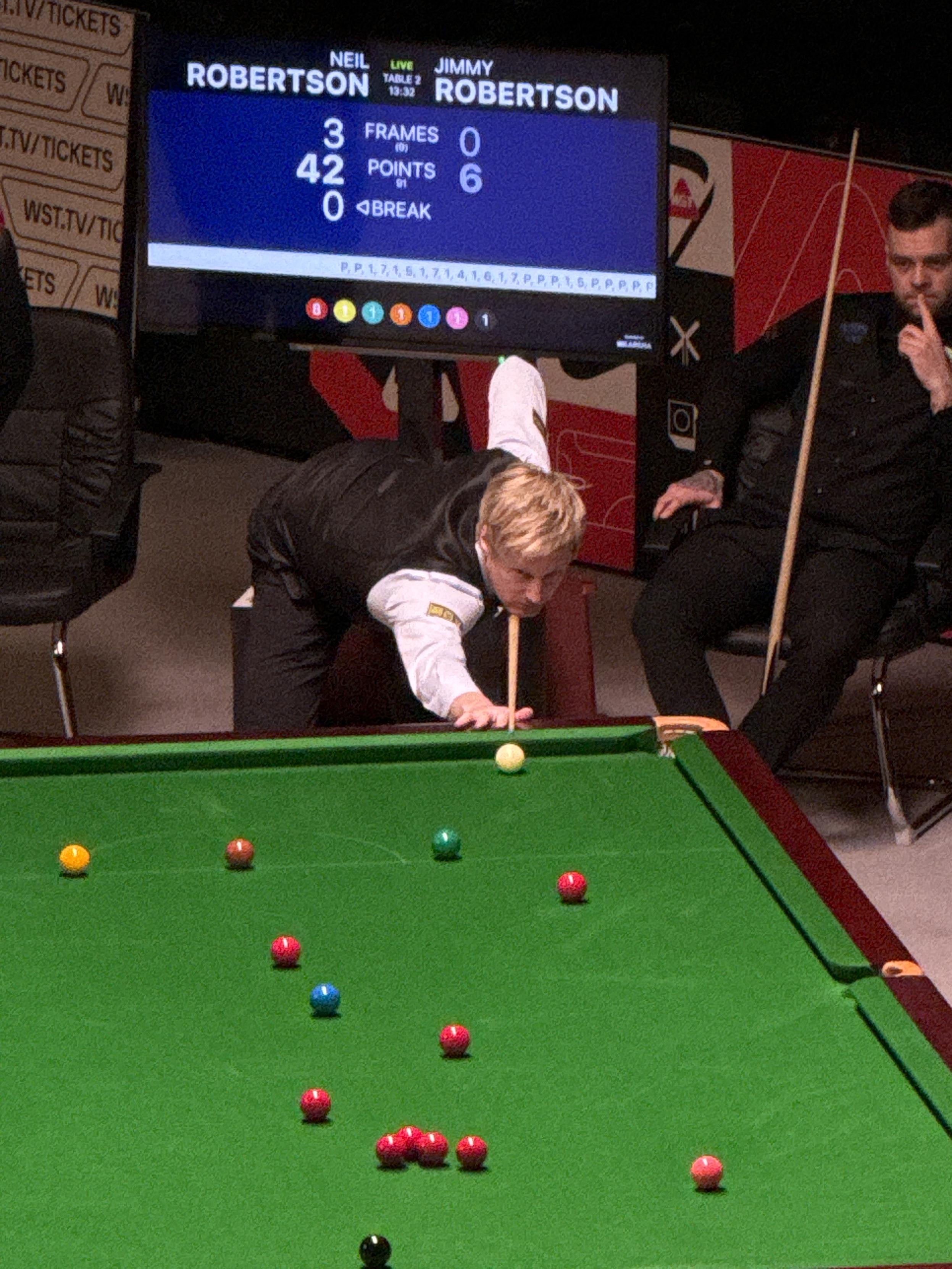 Mecz snookera, ćwierćfinał German Masters. Neil Robertson pochylony do oddania uderzenia w kierunku czerwonych bil. Za nim w fotelu siedzi przyglądający mu się rywal, Jimmy Robertson.