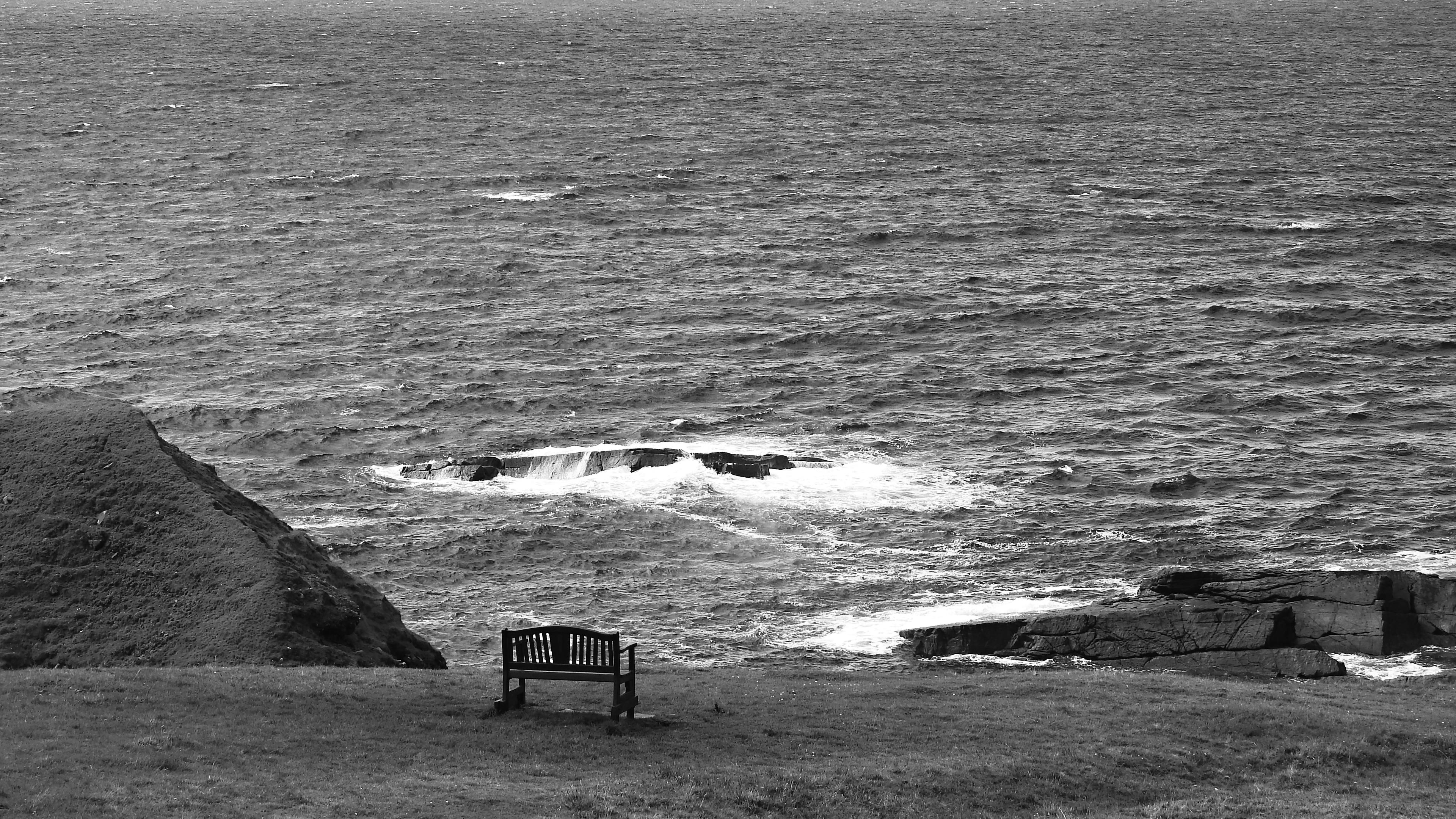 The black and white photograph shows an empty bench on the edge of a cliff in Scotland. Surrounded by sea, greenery and rocks.