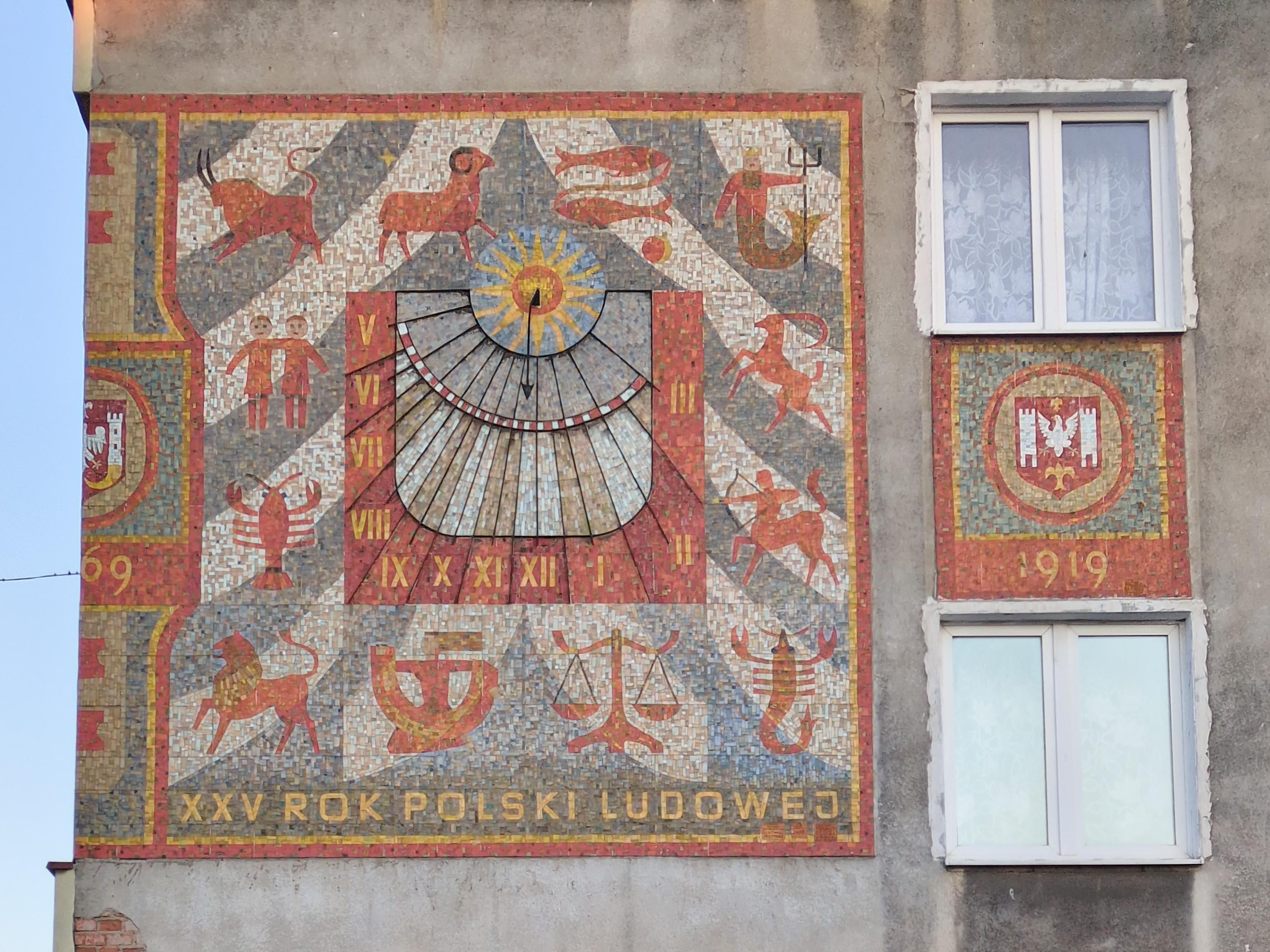 Mural u góry budynku. Dominuje czerwień i żółć na szaro-biało-zielonym tle.

Pośrodku okrągły malunek żółto-pomarańczowego słońca na niebieskim tle z zegarem słonecznym, ukazującym godziny od piątej (rano) do trzeciej (po południu), w postaci nierównych pól, wyciętych po łuku na planie prostokąta, dzielących każdą godzinę na pół. Godziny opisano liczbami rzymskimi koloru żółtego, na czerwonym tle. Pośrodku zegara odchodzi pionowo w dół czarna strzałka, pokazując pierwszą połówkę godziny dwunastej.

Dookoła zegara symbole zodiaku, wykonane na czerwono z żółtymi akcentami, na tle białych i szarozielonych pasów, rozchodzących się ze środka i góry po łukach. U dołu żółty napis na zielonym tle "XXV Rok Polski Ludowej".

Po prawej stronie, pomiędzy dwoma oknami, godło przedstawiające orła (albo kurę?) pomiędzy dwoma wieżami, ze złotymi dynksami u dołu i góry. Pod spodem rok 1919.

Po lewej stronie, na narożniku budynku widać połowę podobnego godła, tyle że tym razem spod wieży do środku odbija jakby złoty róg. Pod spodem widać końcówkę roku "69".