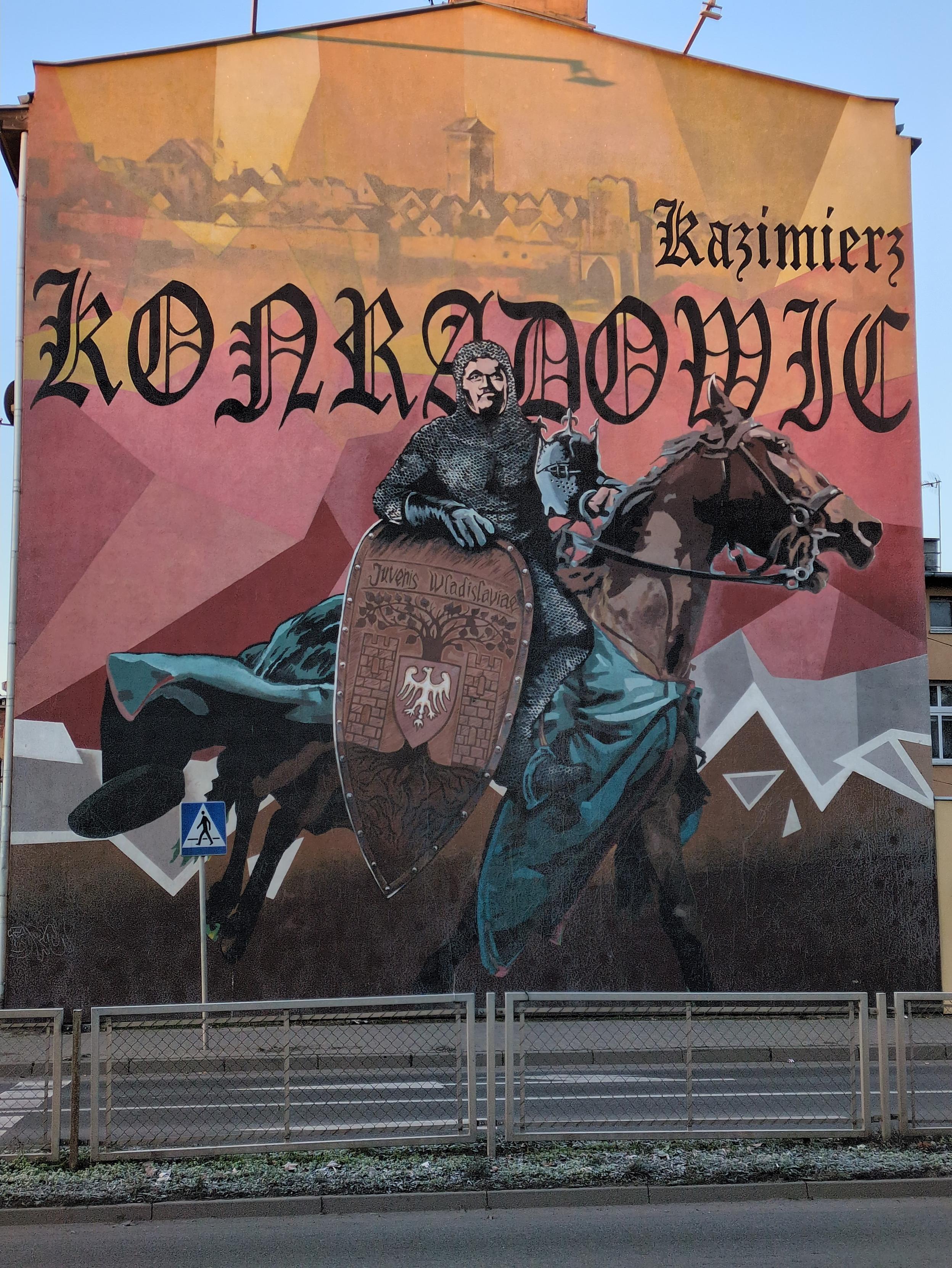 Mural przedstawiający rycerza na kasztanowym koniu. Typ ma na sobie kolczugę, na dłoniach skórzane rękawice, w ręku trzyma (oboj lejc) pełny hełm z trzema szpikulcami na wierzchu. Całe to żelastwo ma lekko zielono-cynowe zabarwienie. Twarz ma nieco kubistyczną, brodatą. Koń biegnie, ma otwarty pysk, zielononiebieską płachtę powiewającą do tyłu. Z boku wysoka, brązowa tarcza z orlim godłem pośrodku; pod spodem drzewo, przedstawione z gęstą siecią korzeni u dołu, wysokim pniem pośrodku i rozprzestrzeniającymi się na boku gałęziami z nieproporcjonalnie wielkimi liśćmi u góry. Obok drzewa dwie kamienne wieże. U góry "Juvenis Wladislaviae". Tło kanciasto-kubistyczne, od spodu brązowe, wyżej szare, czerwone i pomarańczowe. U góry na tle tego pomarańczu kontur panoramy miasta, a na nim ozdobnym krojem "Kazimierz KONRADOWIC"..