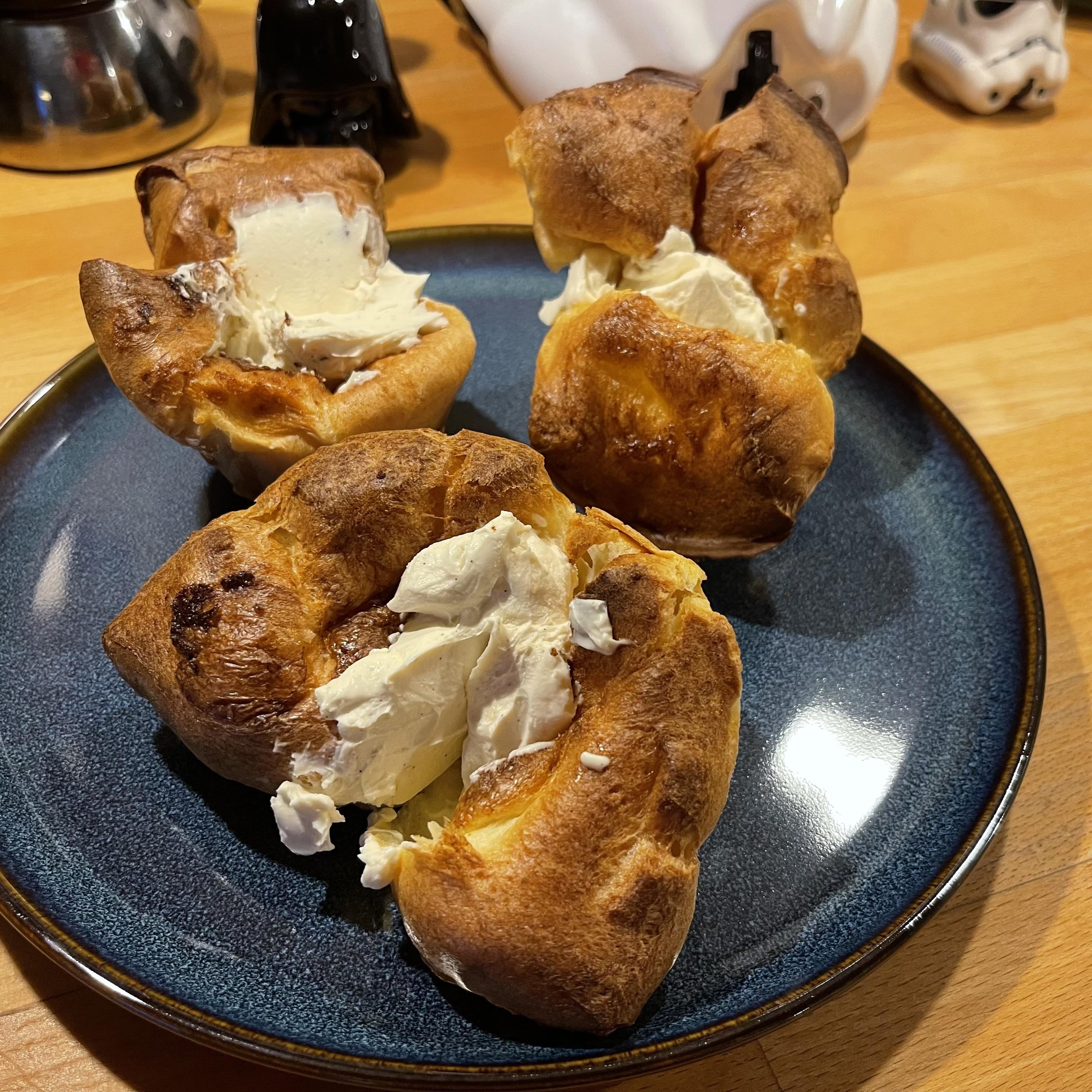 Na granatowym talerzu leży pięć złocistych, pieczonych babeczek z wyrośniętego ciasta, wypełnionych jasnym waniliowym mascarpone. Talerz stoi na drewnianym blacie kuchennym, w tle widoczne są naczynia kuchenne.