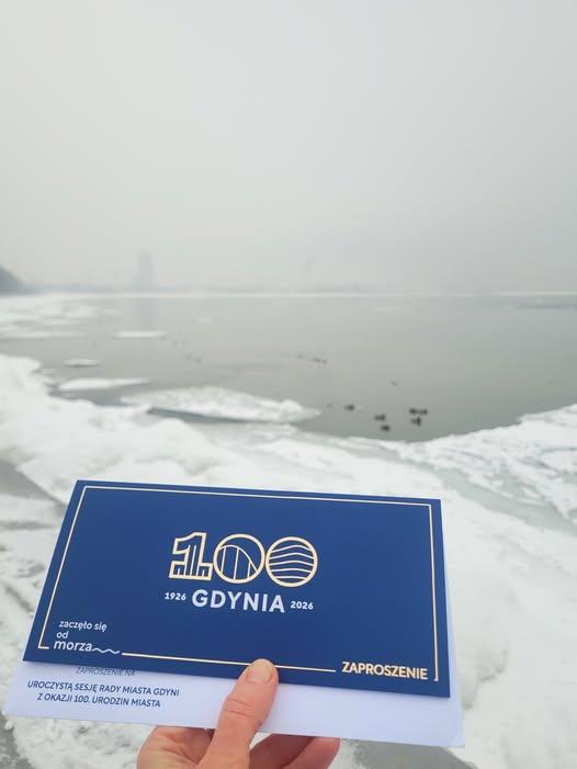 Ręka trzyma granatowe zaproszenie z napisem „100 Gdynia 1926–2026” oraz „Zaproszenie” na tle zimowego, zaśnieżonego brzegu morza z kry i spokojną wodą w oddali.