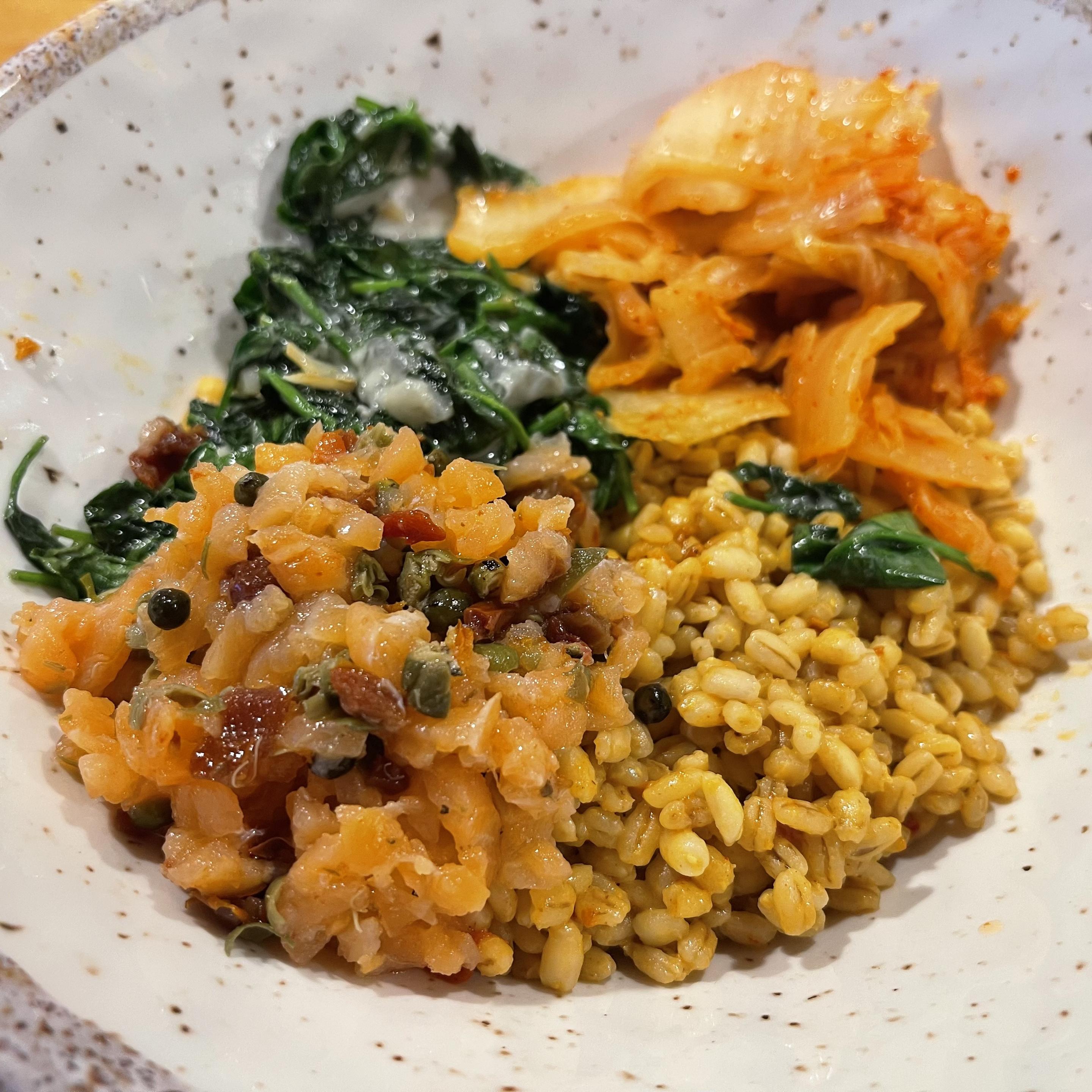 W jasnej, ceramicznej misce znajduje się kompozycja z czterech składników: złocisty pęczak w kremowym sosie, drobno posiekany tatar z łososia z kaparami, blanszowany szpinak z roztopioną gorgonzolą oraz porcja pomarańczowego kimchi. Składniki ułożone są obok siebie, tworząc kontrast kolorów zieleni, pomarańczu i złota.