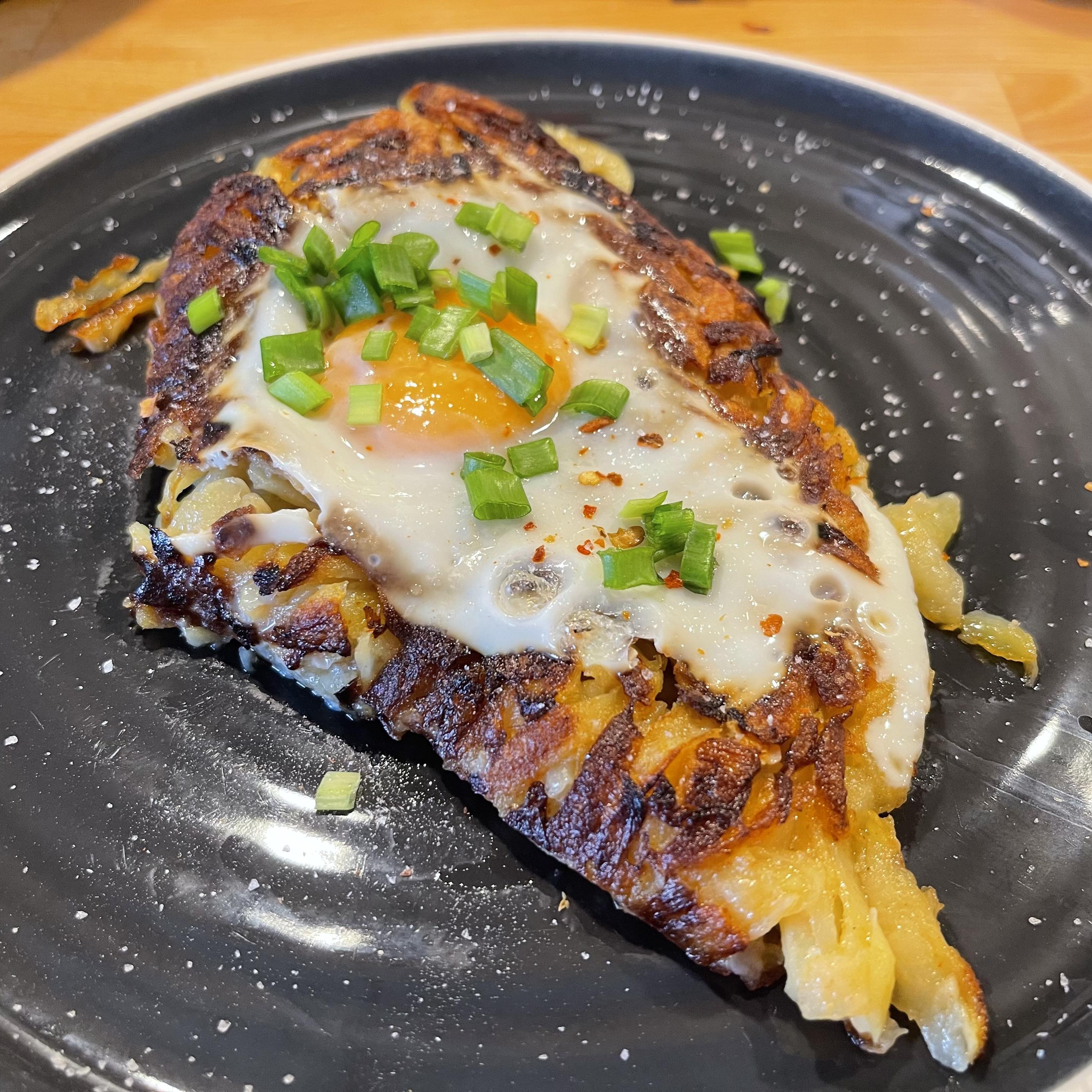 Na czarnym talerzu leży złocisty hash brown z chrupiącą, przypieczoną skórką. Na wierzchu znajduje się jajko sadzone z płynnym żółtkiem.