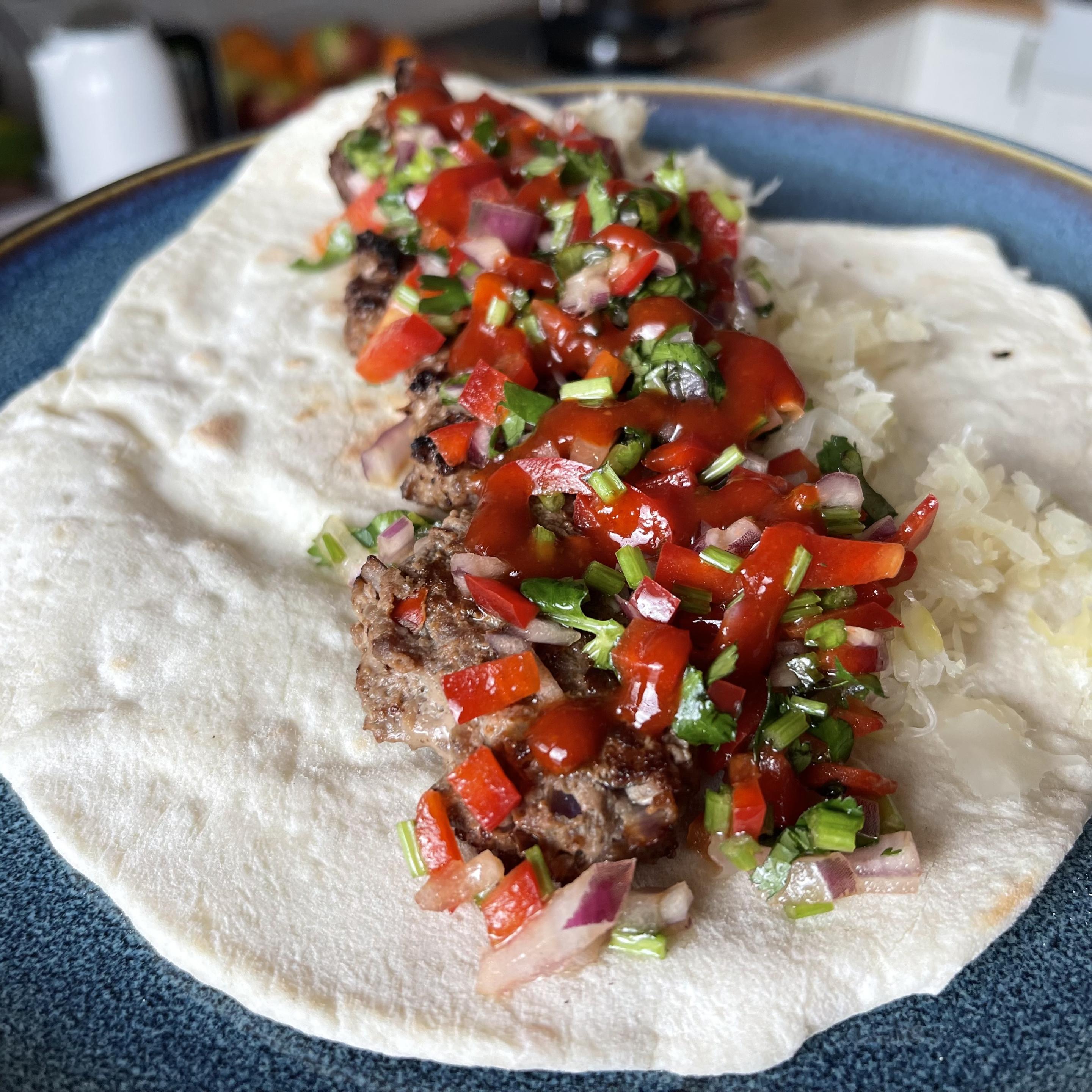 Na niebieskim talerzu leży pszenny podpłomyk wypełniony grillowanymi koftami z wołowiny. Na mięsie znajduje się porcja kiszonej kapusty oraz świeże pico de gallo z czerwonej papryki, czerwonej cebuli i ziół, całość polana czerwonym sosem sriracha.
