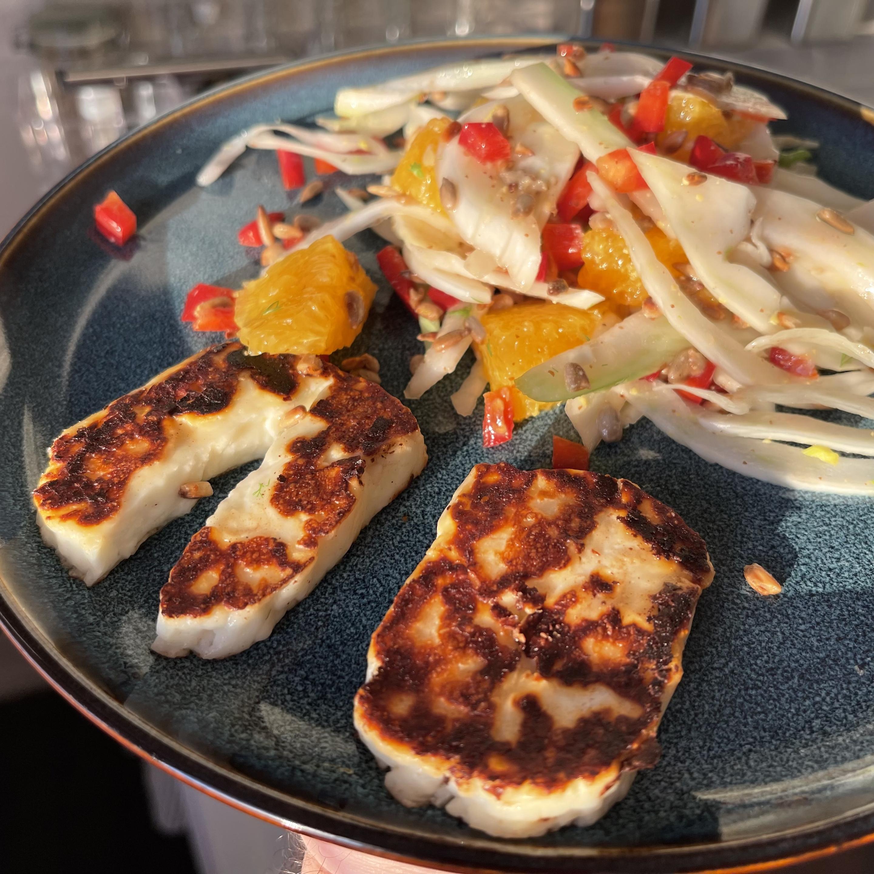 Na ciemnoniebieskim talerzu leżą trzy plastry grillowanego sera halloumi z wyraźnie przypieczoną, złotobrązową skórką. Obok znajduje się sałatka z cienko krojonego fenkuła, kawałków pomarańczy, czerwonej papryki i prażonych ziaren słonecznika
