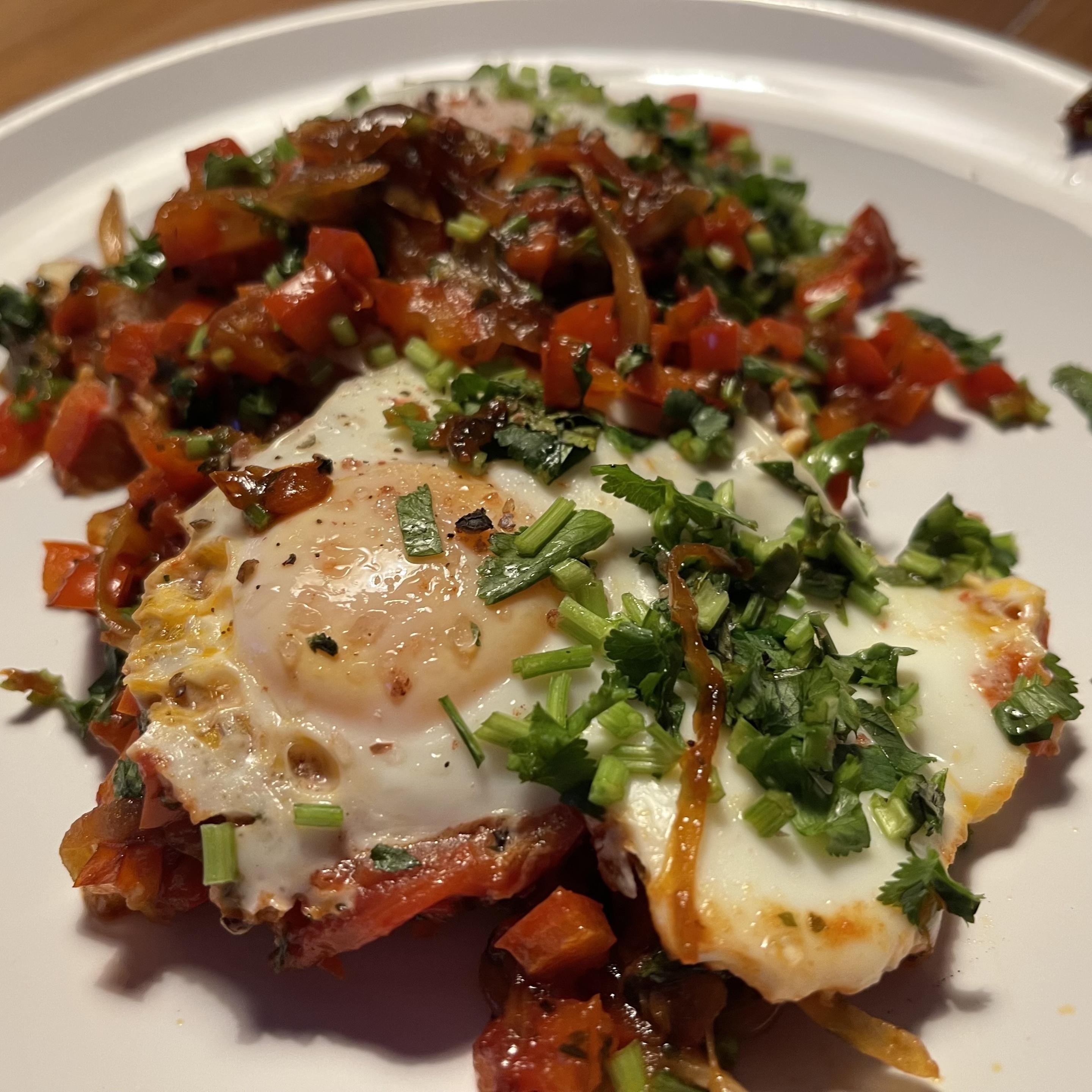 Na białym talerzu znajdują się dwa jajka sadzone podane na duszonej czerwonej papryce i cebuli. Całość posypana jest świeżą kolendrą