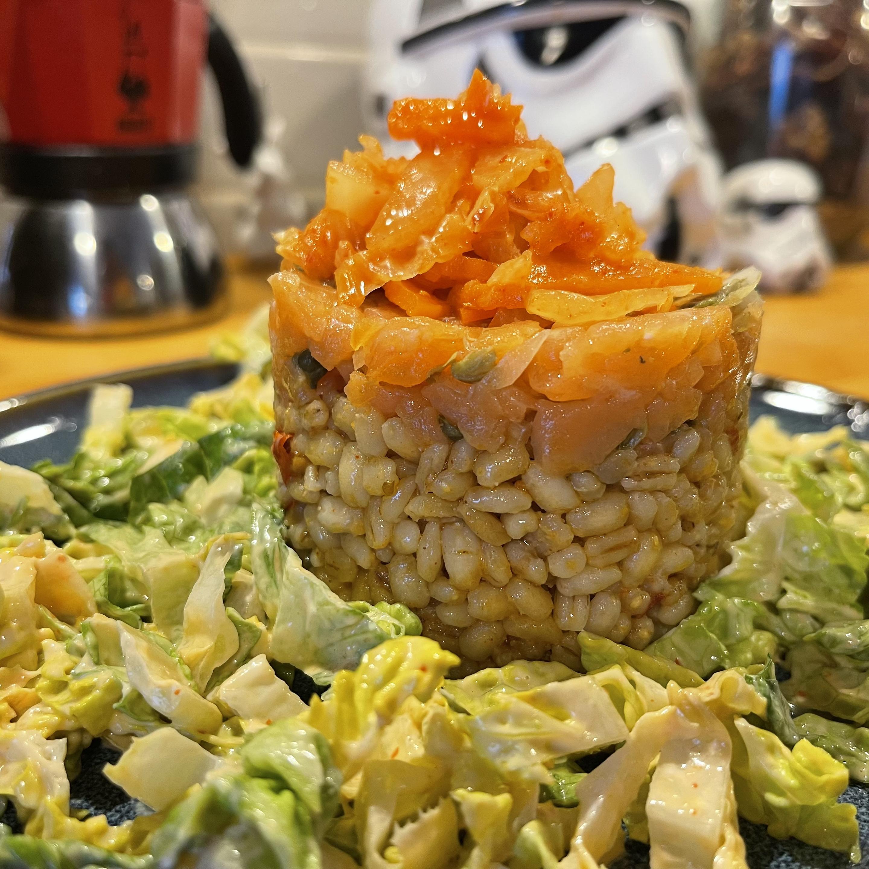 Na talerzu uformowana w cylindryczny kształt kasza pęczak w jasnobrązowym sosie, na wierzchu tatar z łososia z widocznymi ziarnami przypraw i drobno krojonymi dodatkami. Całość otoczona sałatą rzymską w kremowym sosie o lekko pomarańczowym odcieniu. W tle rozmyta kuchnia z czajnikiem i figurką szturmowca.