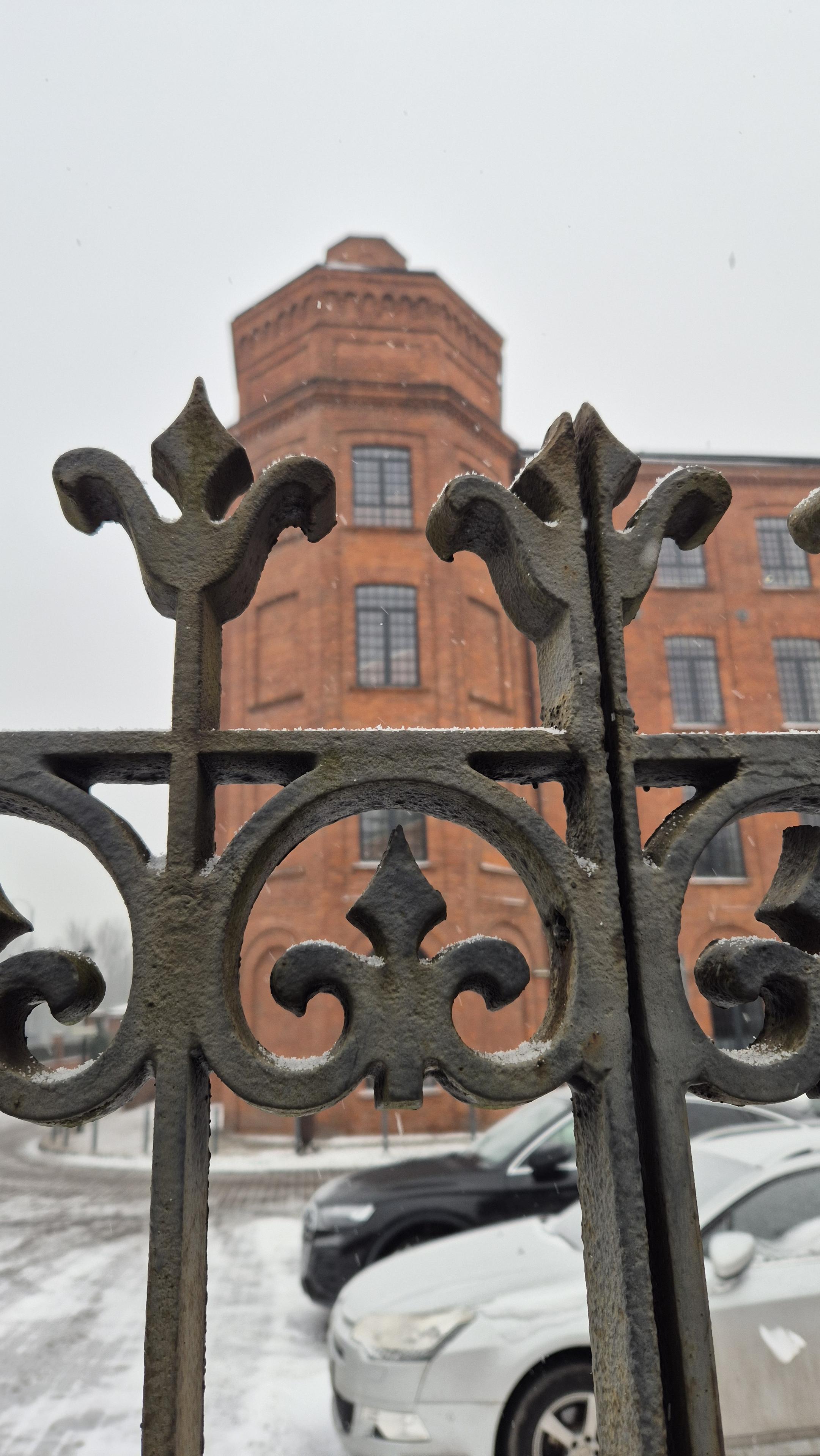 Detal ogrodzenia wspomnianego budynku. W tle narożna wieża klatki schodowej, imitująca stylem wieże zamkowe.