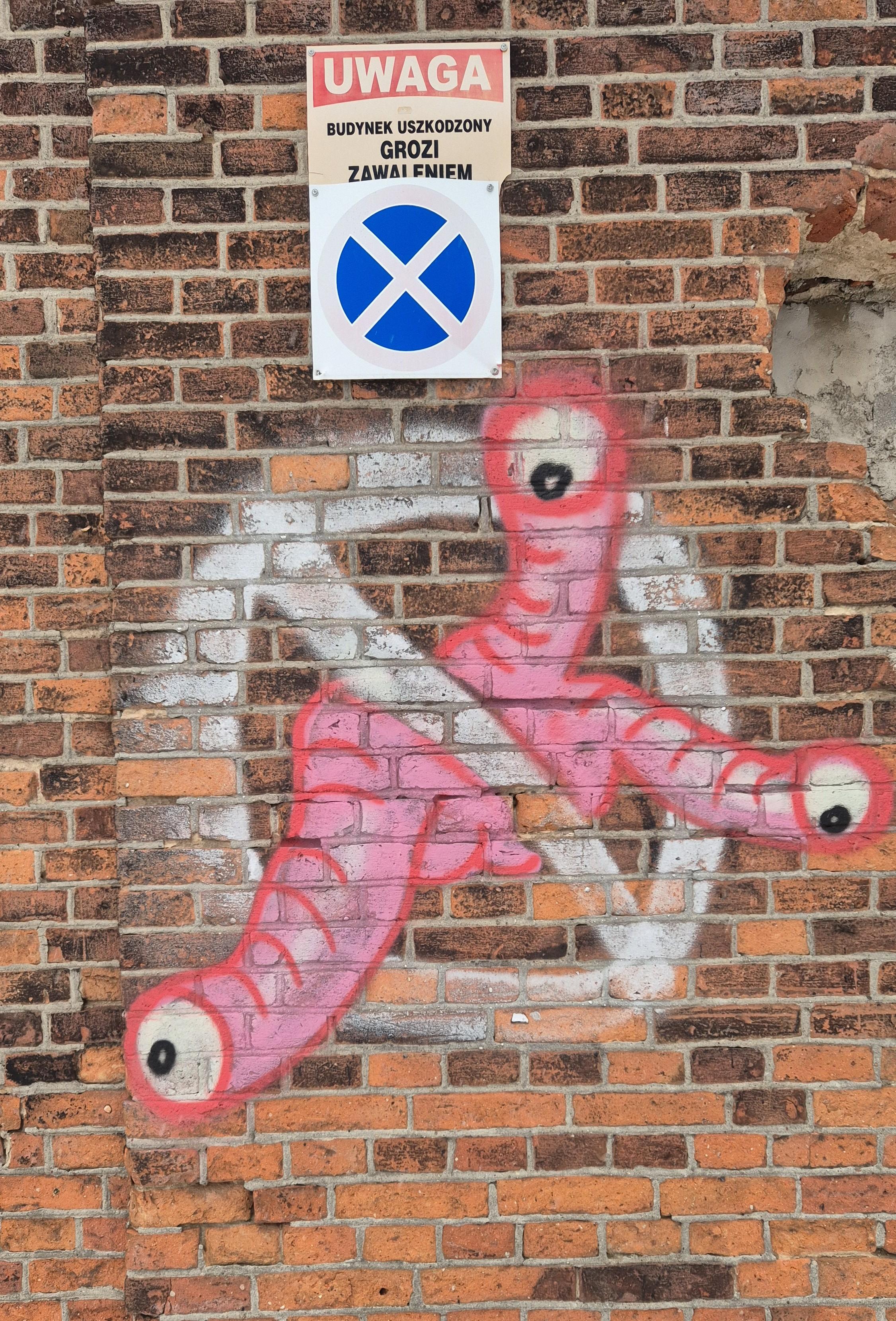 Ceglana ściana z graffiti przedstawiającym różowego potwora z wieloma oczami, podobnego do kłębowiska dżdżownic. Potwór jest przekreślony znakiem zakazu. Przywodzi na myśl Shoggothy z opowiadań H.P.Lovecrafta.