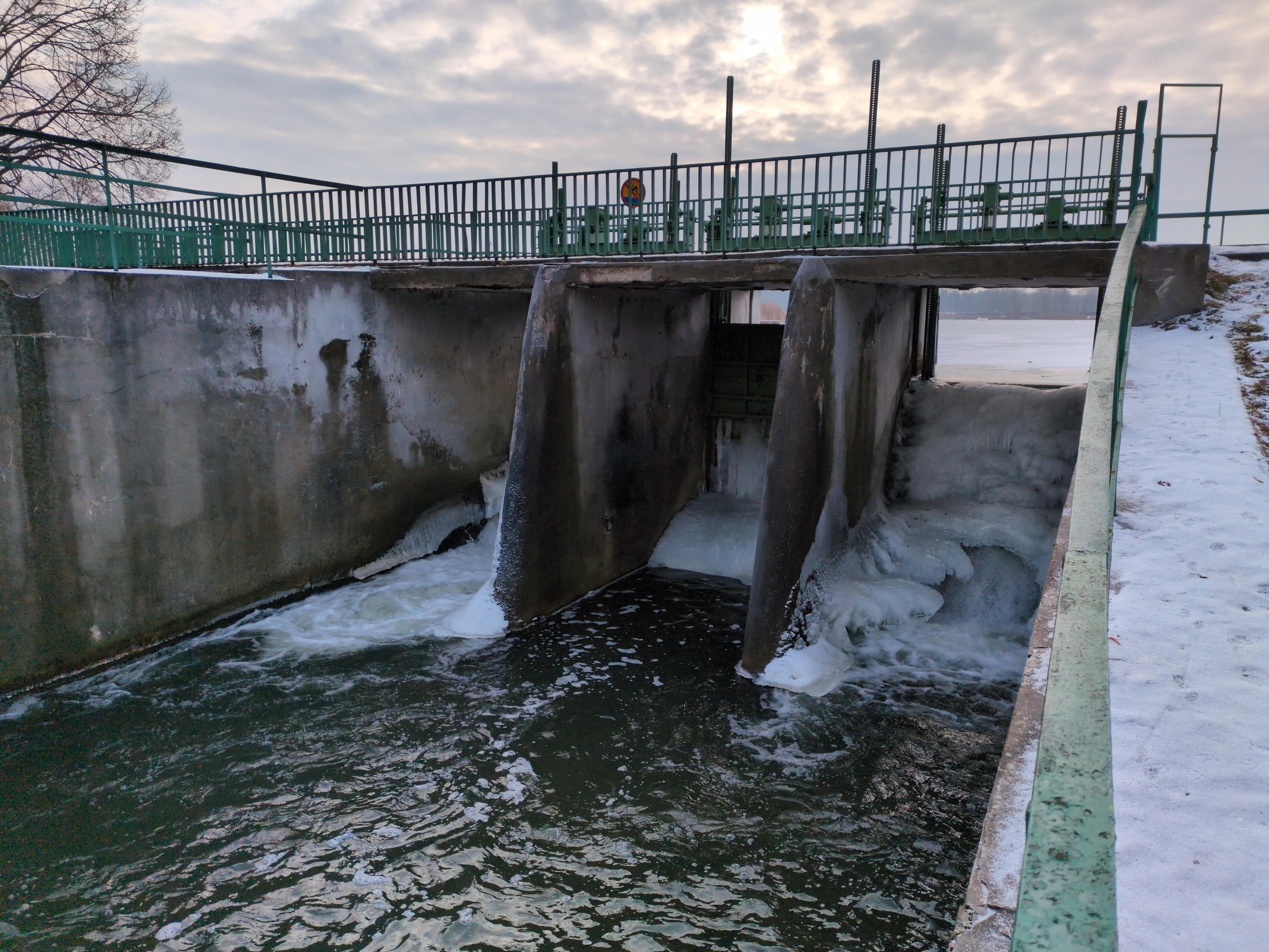 Jaz na rzece, widziany pod katem z dolnej strony. Podzielony na trzy części szerokimi, betonowymi murami. Środkowa przegroda jest zasunięta, poniżej metalowej zasłony widać zamarzniętą pionową ścianę wody, a poniżej niej zamarznięty łuk. Przez dwie pozostałe przegrody przepływa wzburzona woda, przeciskając się pomiędzy grubymi bryłami lodu o fantazyjnych kształtach, przyklejonymi do ścian jazu. Przez otwór w prawej przegrodzie roztacza się widok na zamarznięte jezioro, a na horyzoncie las. Ze względu na kąt zdjęcia końca lewej przegrody nie widać. Na jazie kładka z zieloną barierką. Woda u dołu wzburzona, w kolorze morskim.