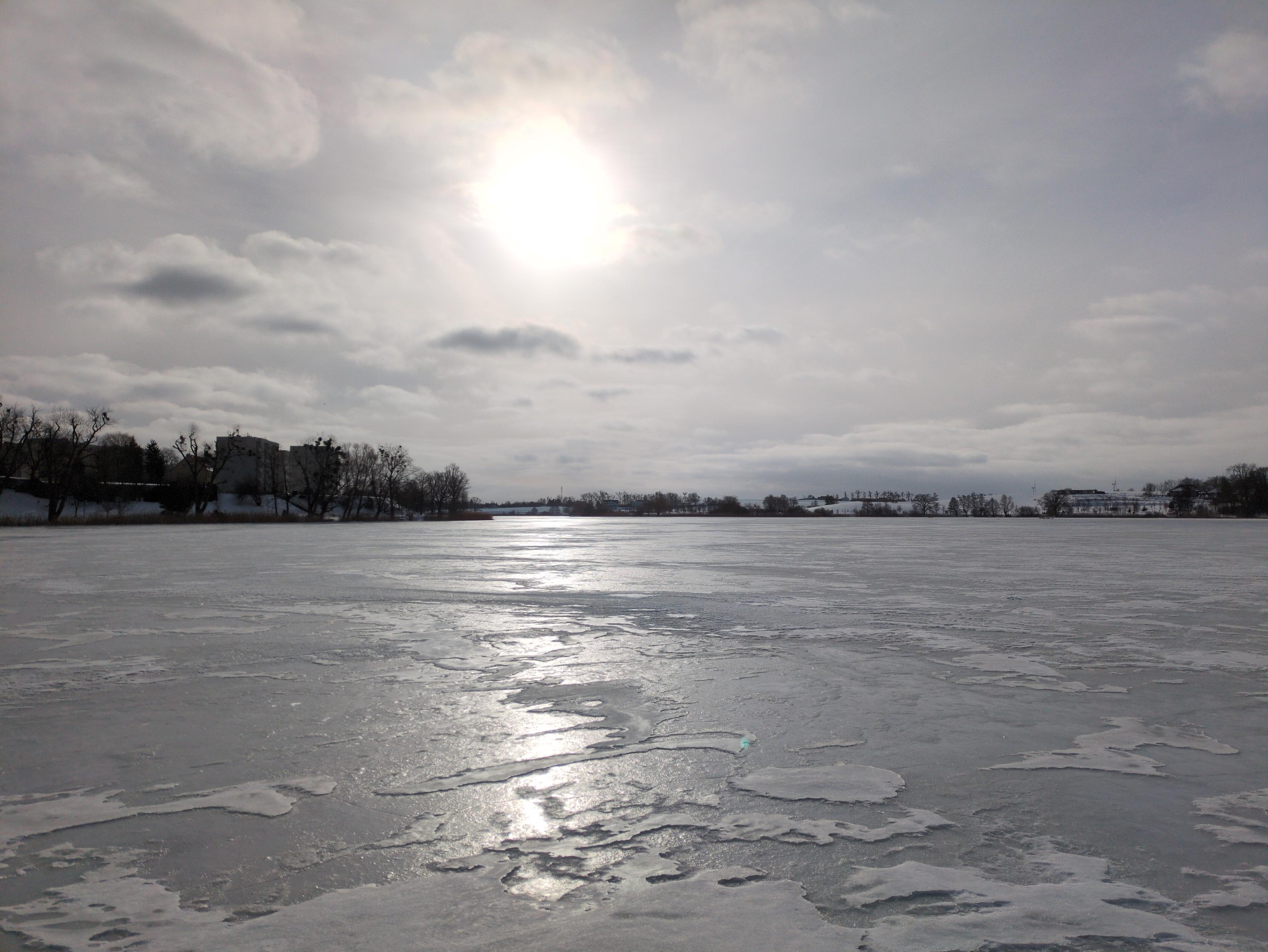 Zamarznięta tafla jeziora w słońcu. Spodnia warstwa lodu tworzy nieco chropowatą, srebrną powierzchnię, na której wystają różnokształtne placki jaśniejszego lodu z wierzchu. Na wprost świeci słońce, tworząc złocisty pas na spodniej warstwie, przerywany plackami warstwy wierzchniej. Niebo również srebrzyste, z kilkoma ciemniejszymi chmurami. Na brzegu jeziora widać pojedyncze drzewa, a dalej niewyraźne zabudowania; bliżej po stronie lewej, a dalej na horyzoncie. Nad prawą stroną horyzontu ciągła warstwa białych chmur.