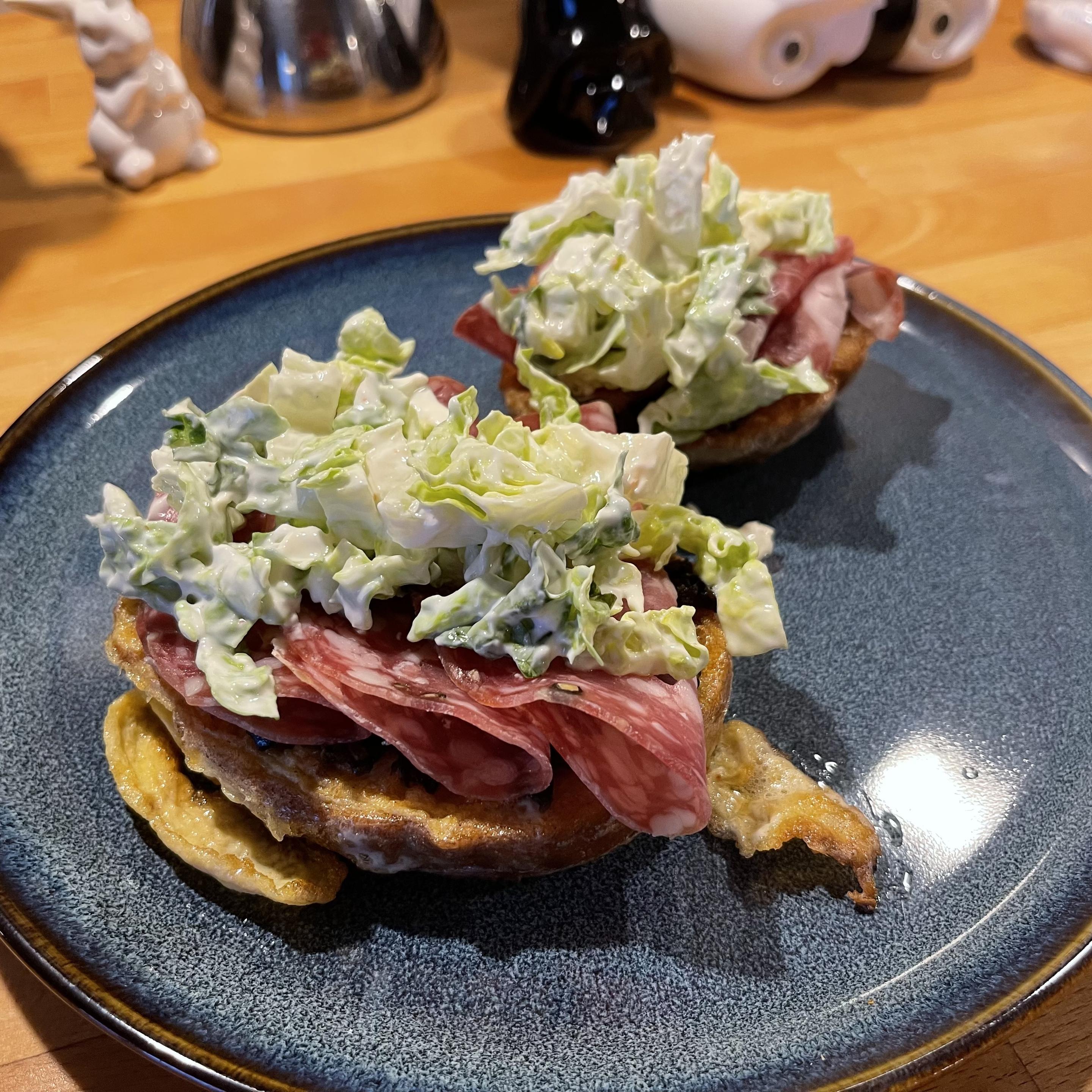 Na ciemnym talerzu leżą dwa grube tosty francuskie. Na każdym ułożone są plastry salami, a na wierzchu porcja sałaty rzymskiej w jasnym, kremowym sosie jogurtowym z widocznymi kawałkami sera pleśniowego. Tosty są złociste i lekko chrupiące na brzegach.