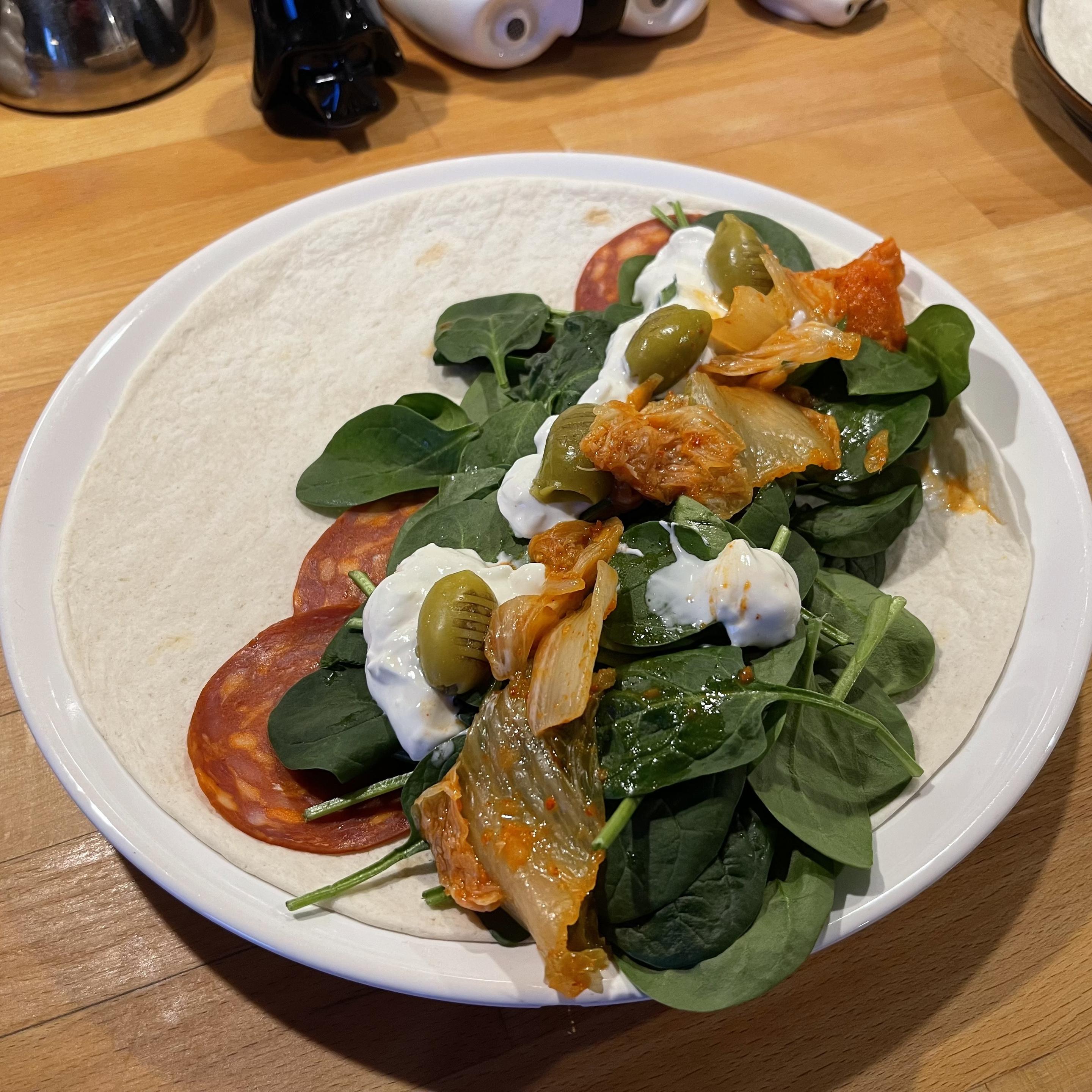 Na białym talerzu leży pszenny placek tortilli. Na jednej połowie ułożone są plastry chorizo, świeże liście szpinaku, kimchi, zielone oliwki oraz kleksy gęstego dipu jogurtowo-gorgonzolowego. Talerz stoi na drewnianym blacie.
