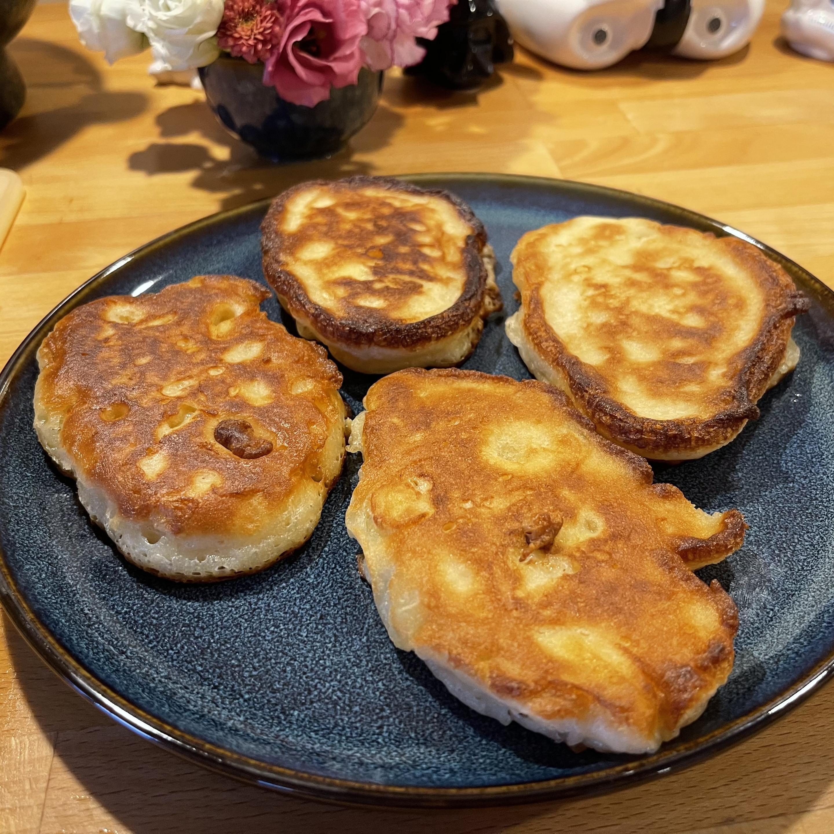 Na ciemnym talerzu leżą cztery rumiane racuszki drożdżowe z widocznymi kawałkami jabłek. Mają złocistą, miejscami mocniej przyrumienioną skórkę i puszyste wnętrze. Talerz stoi na drewnianym blacie, w tle widać niewyraźny bukiet kwiatów.