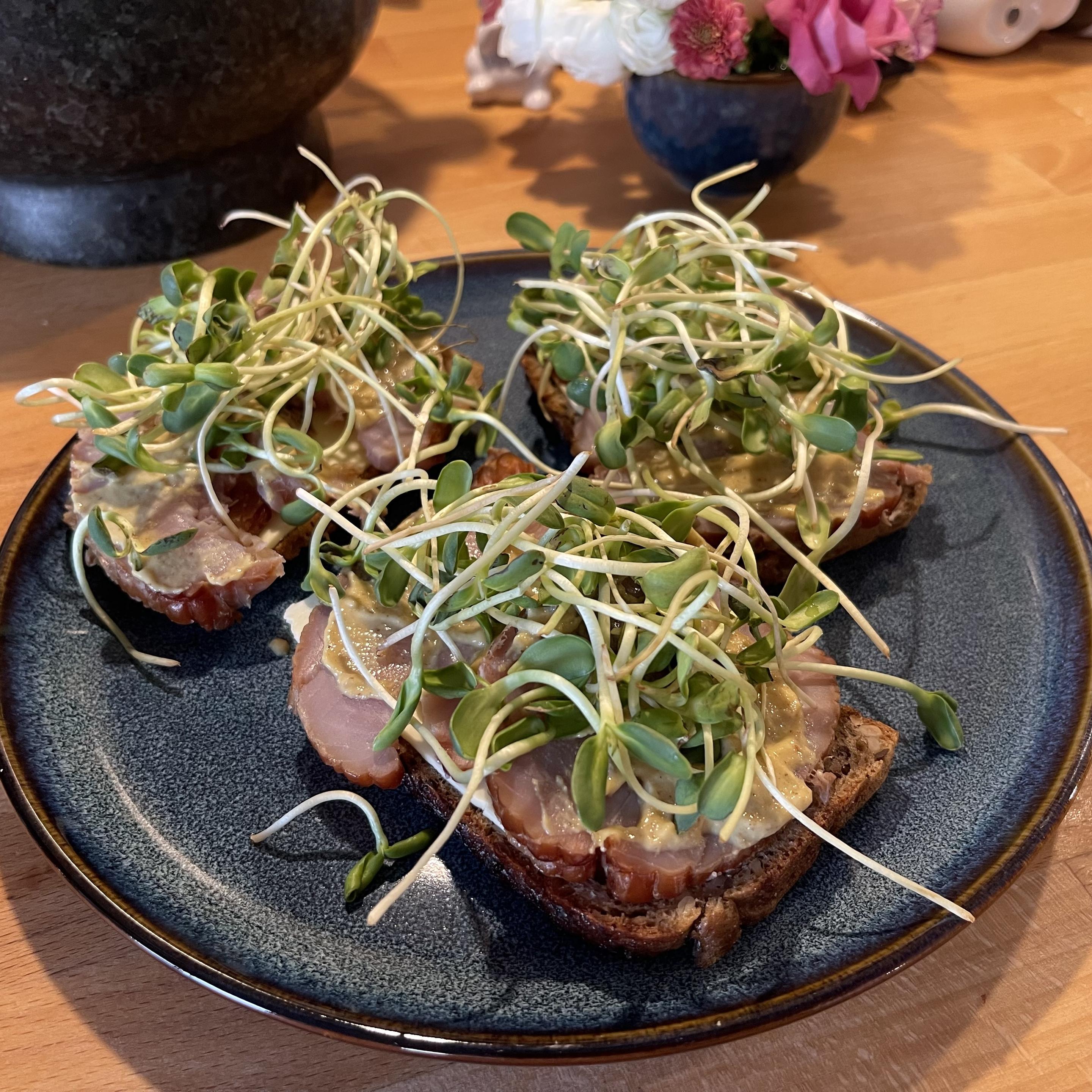 Na granatowym talerzu leżą trzy kromki ciemnego chleba. Każda posmarowana jest sosem musztardowym, przykryta plastrami wędzonej ryby i obficie udekorowana świeżymi kiełkami słonecznika. W tle widać drewniany blat oraz rozmyte kwiaty