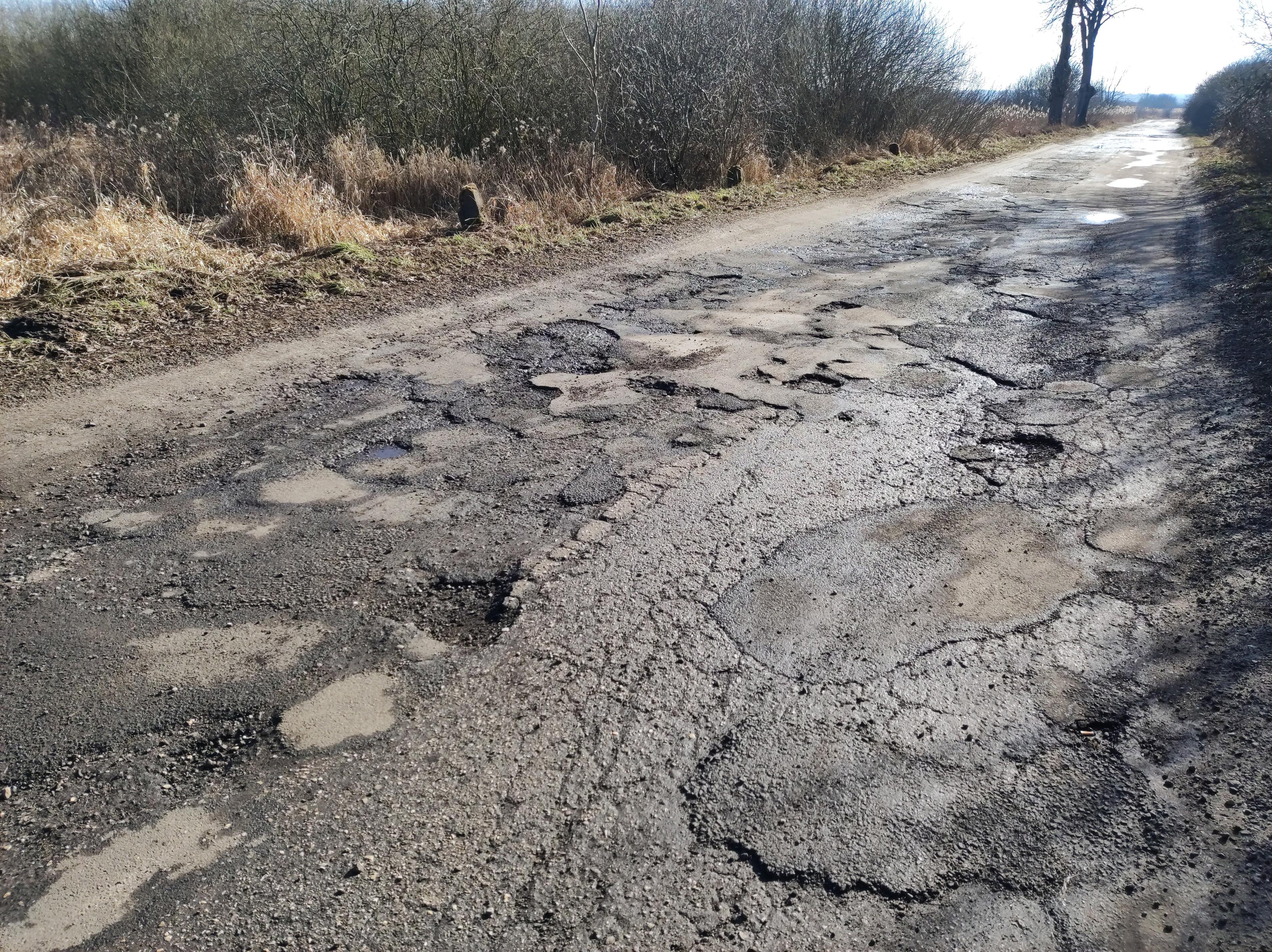 Bardzo zniszczona asfaltowa droga, pełna pęknięć, dziur i łat. Po bokach bezlistne krzewy i sucha trawa.