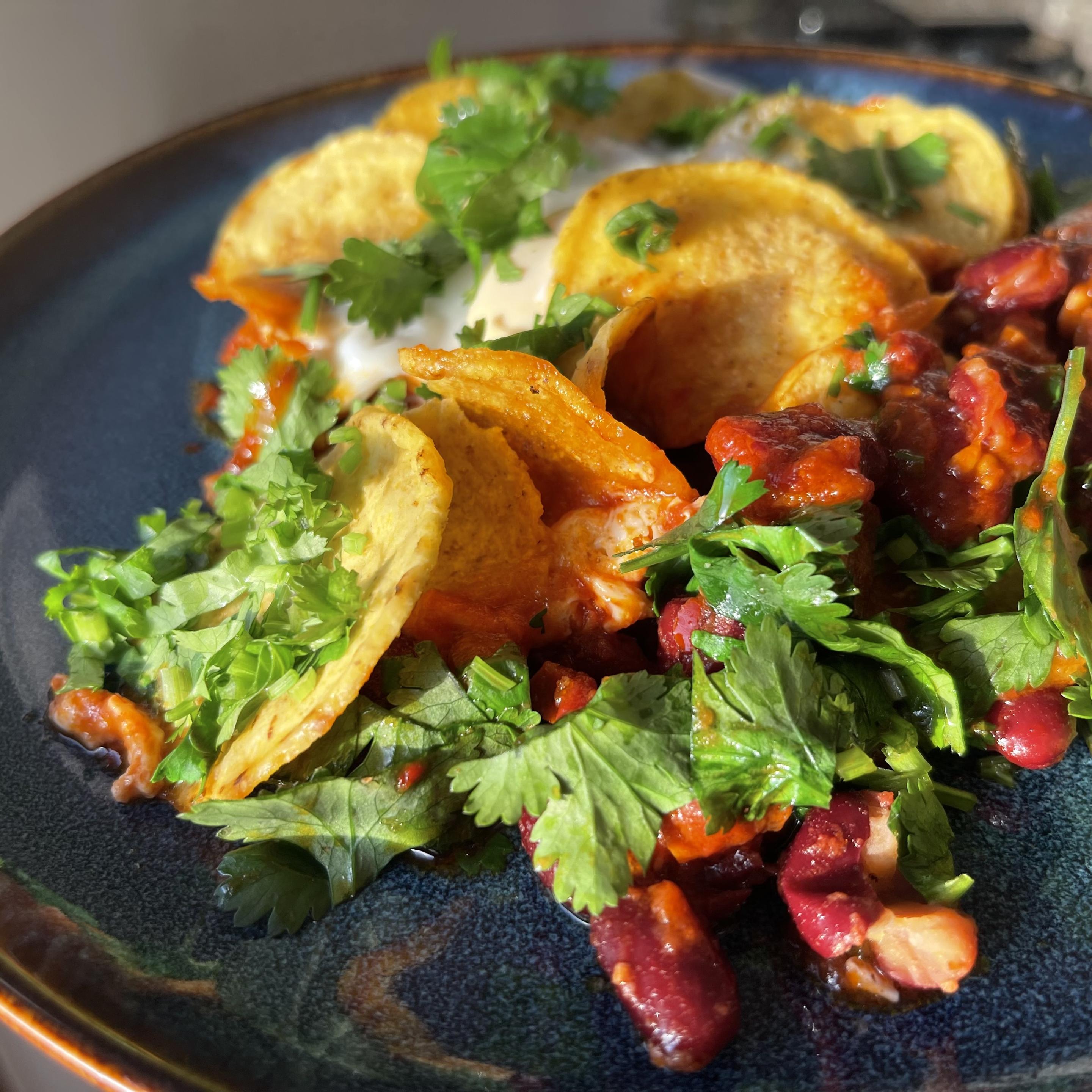 Na granatowym talerzu podane są chrupiące chipsy tortilli pokryte roztopionym serem, czerwoną fasolą w gęstym sosie pomidorowym oraz dużą ilością świeżej kolendry i szczypiorku. Danie jest kolorowe – dominuje złoto chipsów, czerwień fasoli i intensywna zieleń ziół.