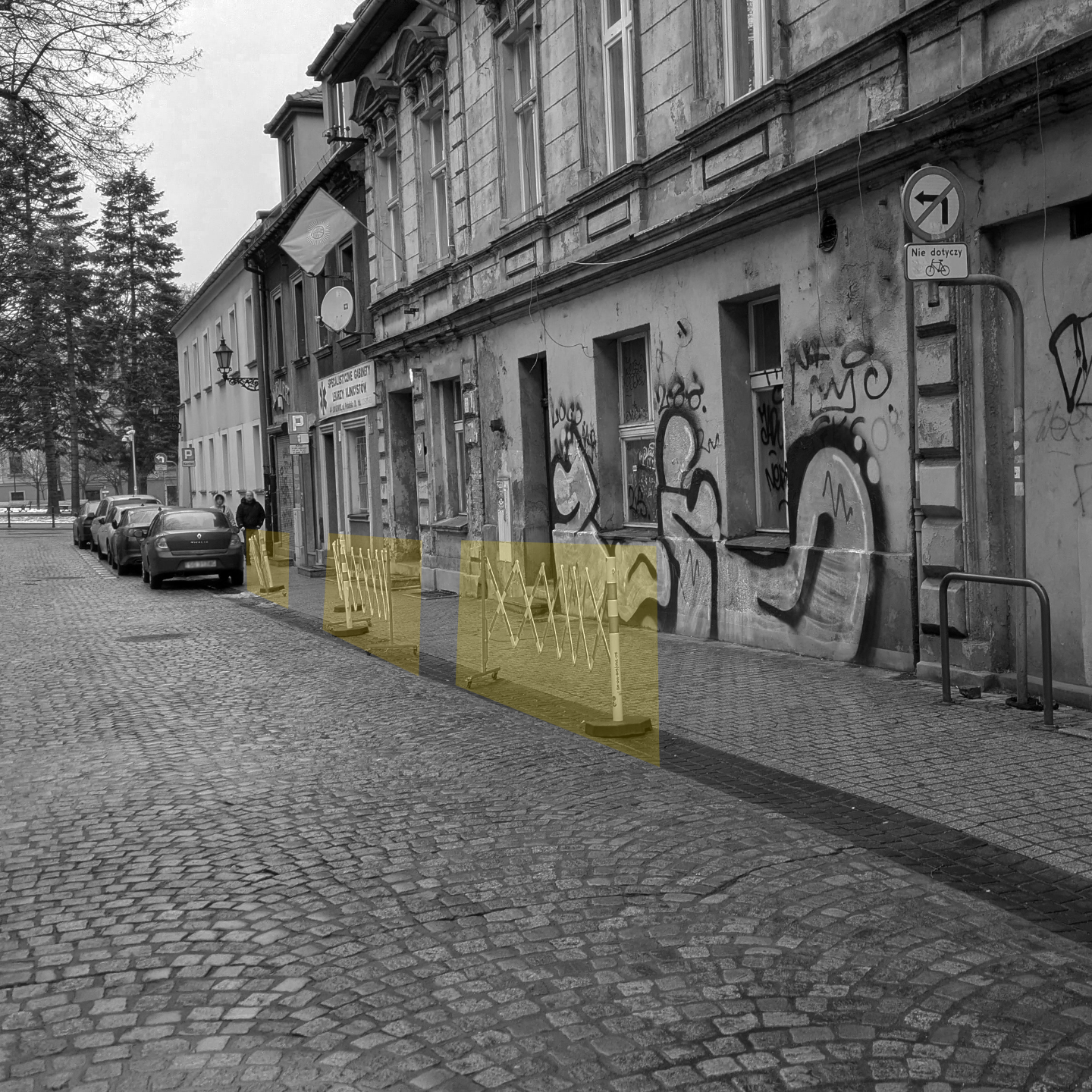 Czarno białe zdjęcie uliczki starego miasta, na żółto zaznaczone rozkładane barierki blokujące pojazdom parkowanie.