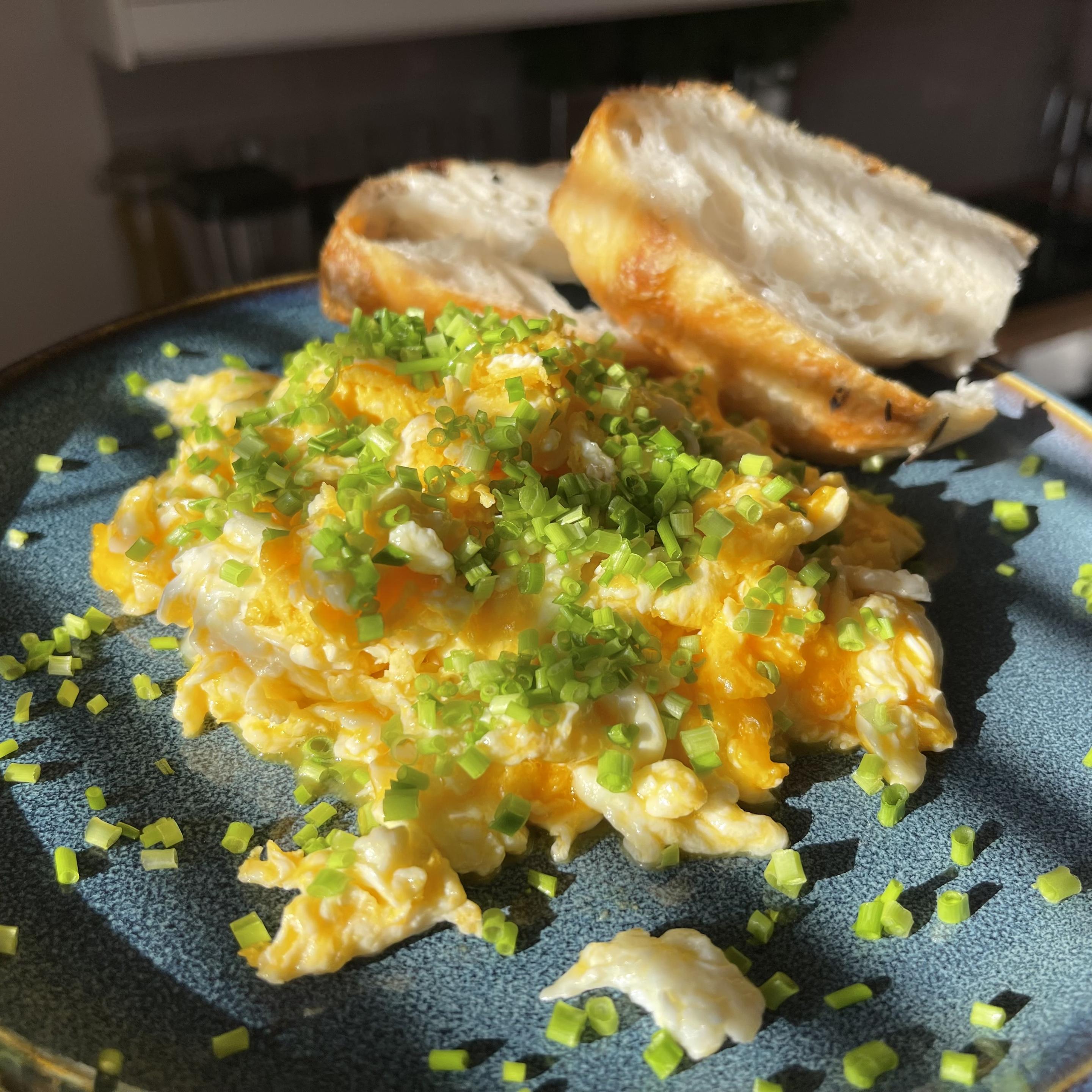 Na ciemnym talerzu znajduje się porcja kremowej jajecznicy z widocznymi kawałkami ściętego białka i płynnego żółtka, posypana dużą ilością świeżego szczypiorku. Z tyłu leżą dwie połówki domowej focacci o puszystym wnętrzu i złocistej skórce. Całość oświetlona jest ciepłym światłem.