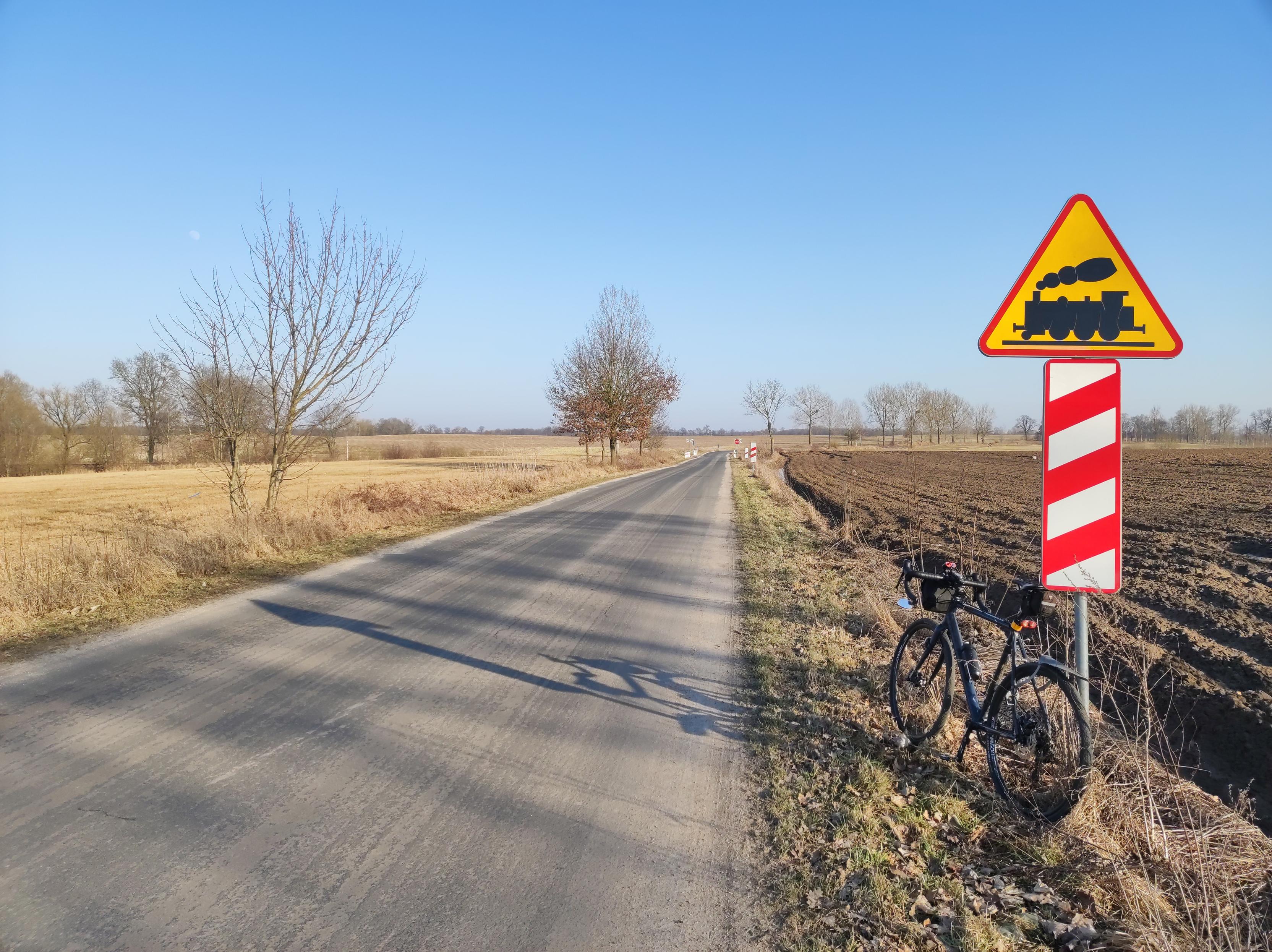 Przy nieduża asfaltowa szosie stoi znak ostrzegający o przejeździe kolejowym, o który opiera się tylnym kołem ciemnoniebieski rower szutrowy. Trochę dalej widać przejazd. Niebo jest bezchmurne i błękitne, a dzień słoneczny.