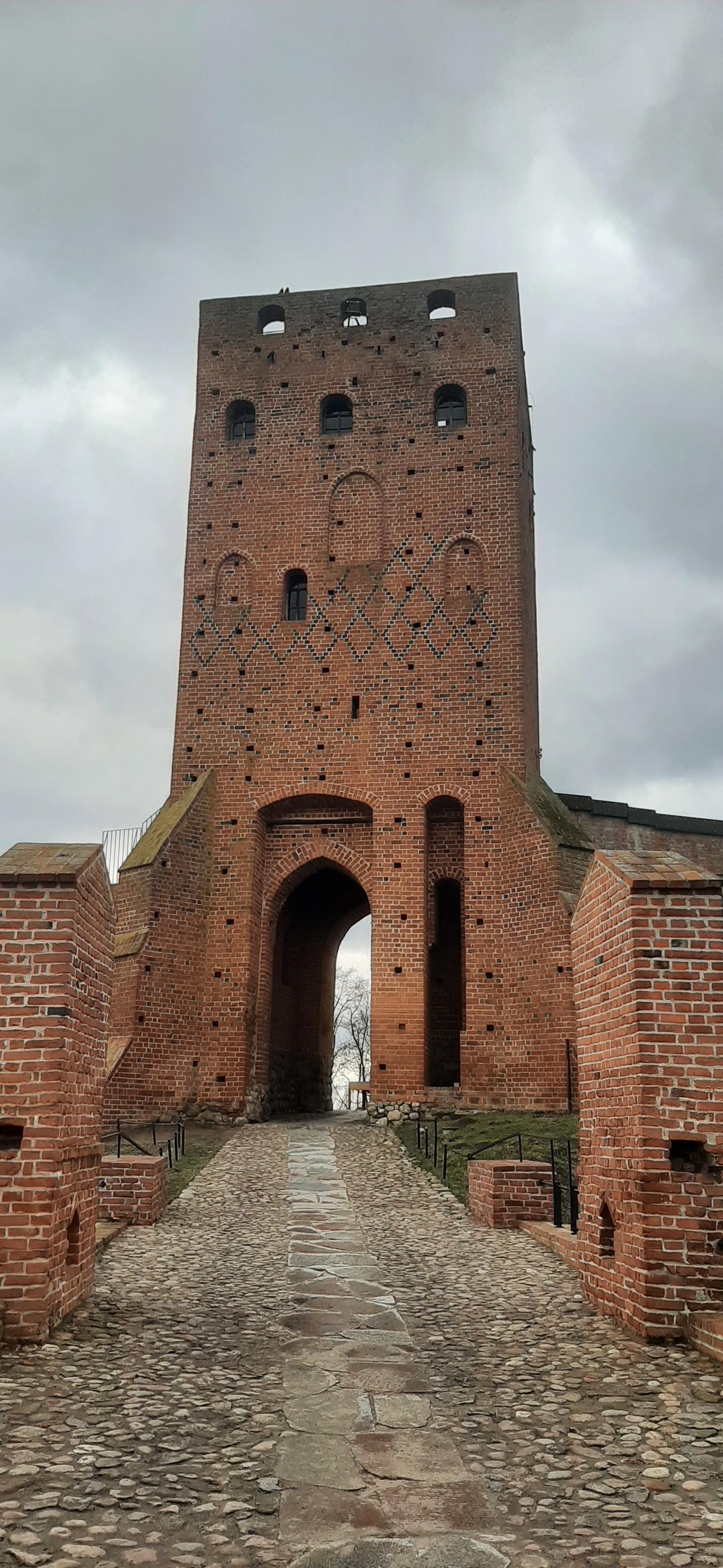 Szeroka, prostopadłościenna gotycka wierza z czerwonej cegły z wysoką, majestatyczną bramą i furtą.