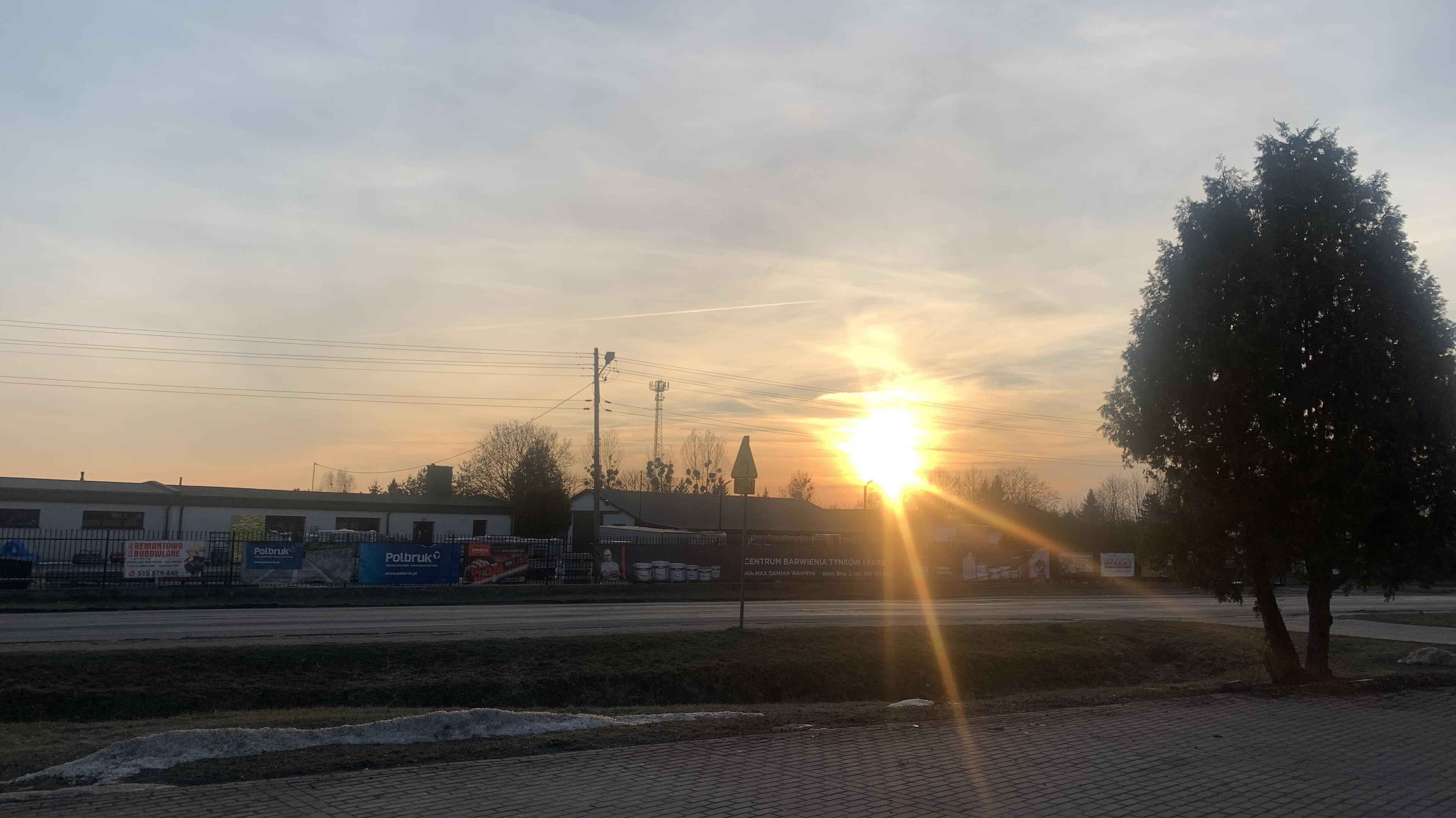 wybrukowany parking, dalej droga i reklama Polbruk na płocie składu budowlanego w gminie. W tle zachodzące słońce