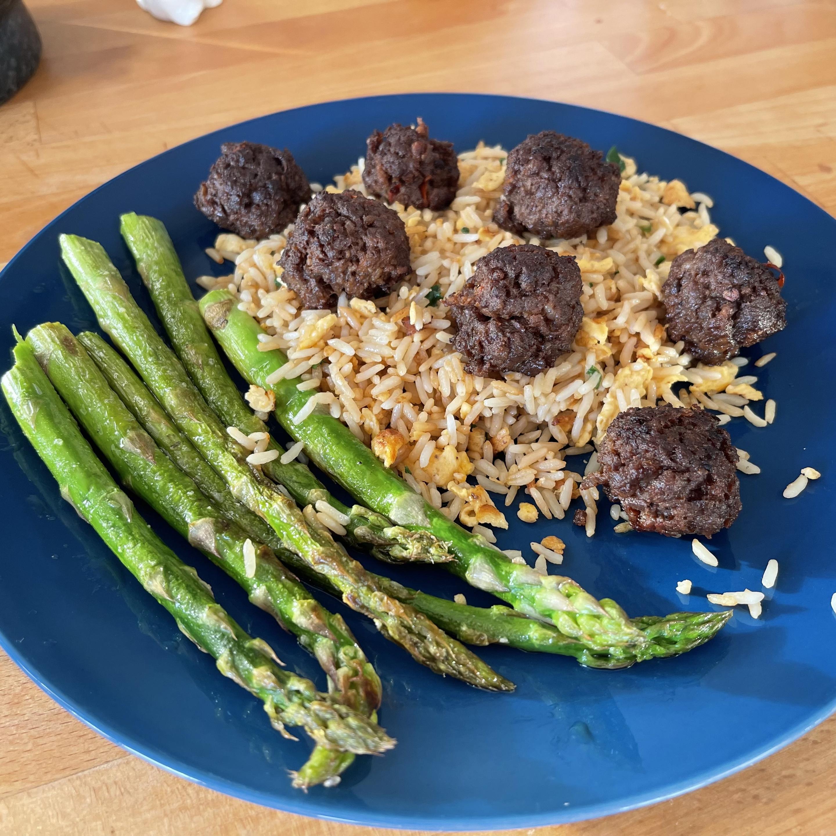 Na niebieskim talerzu znajduje się porcja smażonego ryżu z widocznymi kawałkami jajka. Na wierzchu ułożone są małe, okrągłe pulpeciki wołowe. Obok leży kilka zielonych, podsmażonych szparagów. Talerz stoi na drewnianym blacie.