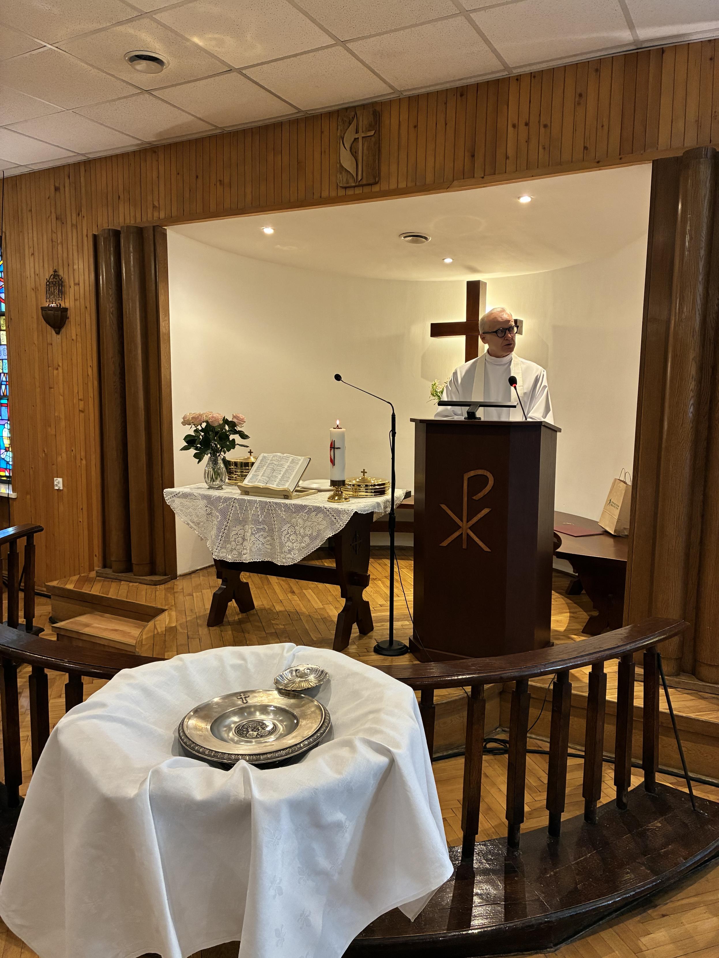Ujęcie na prezbiterium zza stojącej na pierwszym planie chrzcielnicy. Chrzcielnica jest wyłożona białą tkaniną i są w niej ułożone zdobiona, srebrna podkładka oraz małe, płaskie naczynie z wodą. Z tyłu, za półkolistą barierką, stoi nakryty białym obrusem stół z otwartą Biblią, naczyniami liturgicznymi i zapaloną świecą, a obok kazalnica, zza której przemawia mężczyzna w białej albie.