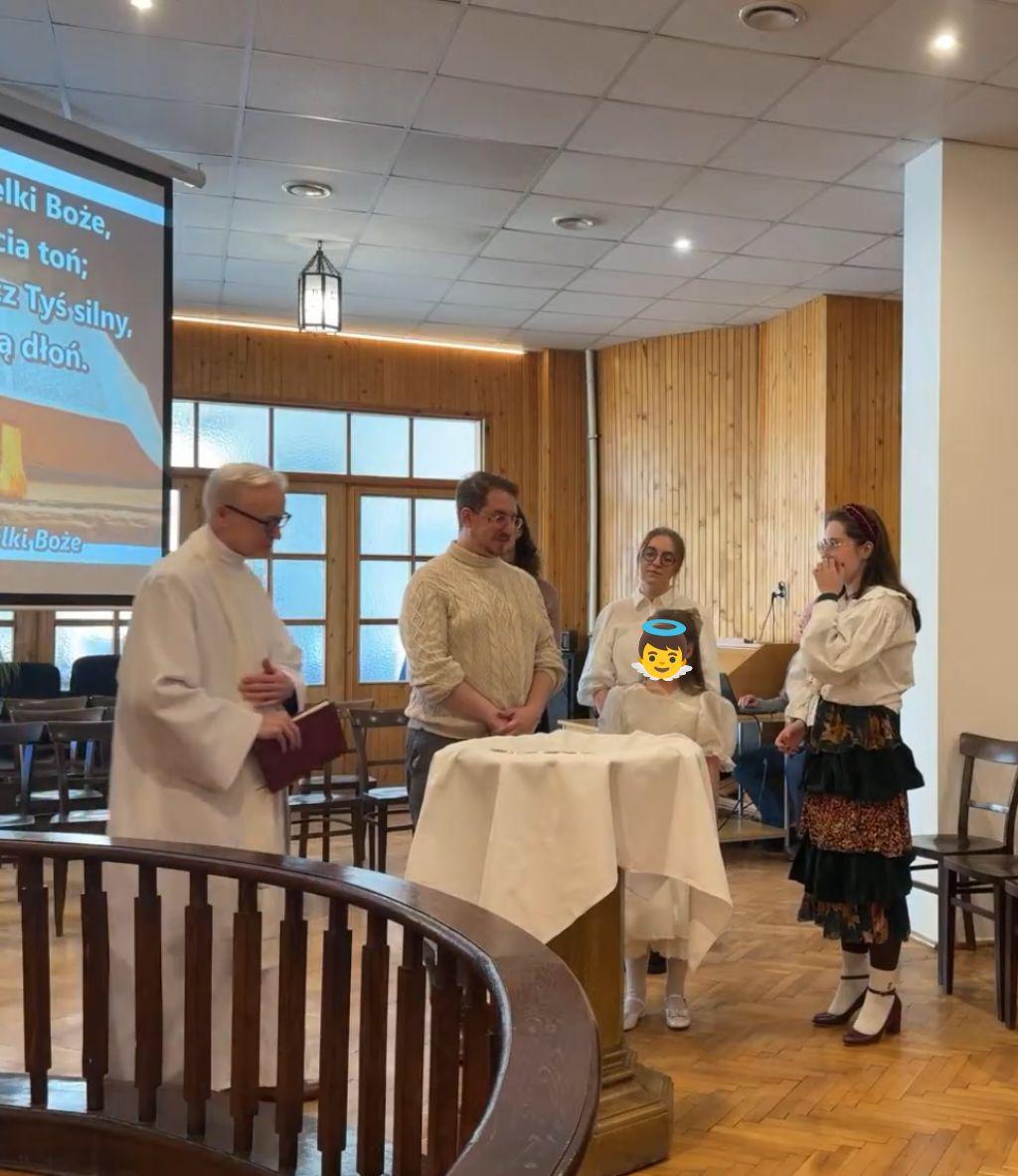 Ujęcie z boku. Przy chrzcielnicy stoi dziewczynka w uroczystej, białej sukience i kilkoro dorosłych. Mężczyzna w albie wyszedł zza barierek i przechodzi obok nich z księgą w dłoni.