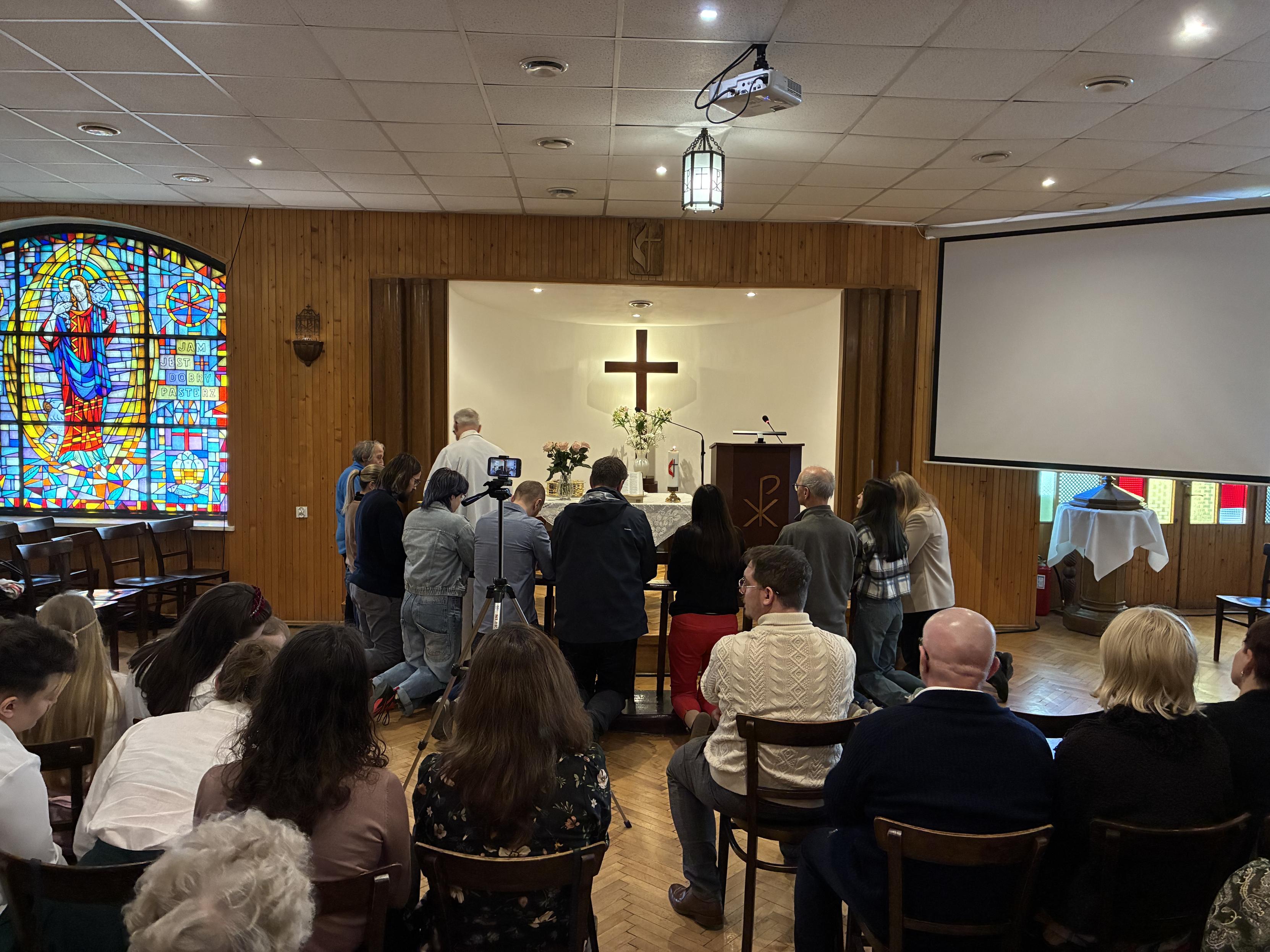 Ujęcie zza pleców siedzących na krzesłach ludzi. Grupa osób klęczy przy barierkach otaczających prezbiterium, z tyłu którego wznosi się podświetlony krzyż, zajmujący centralne miejsce na zdjęciu. Po lewej widać kolorowy witraż z Chrystusem – Dobrym Pasterzem, z prawej już zamkniętą, odsuniętą pod ścianę chrzcielnicę.