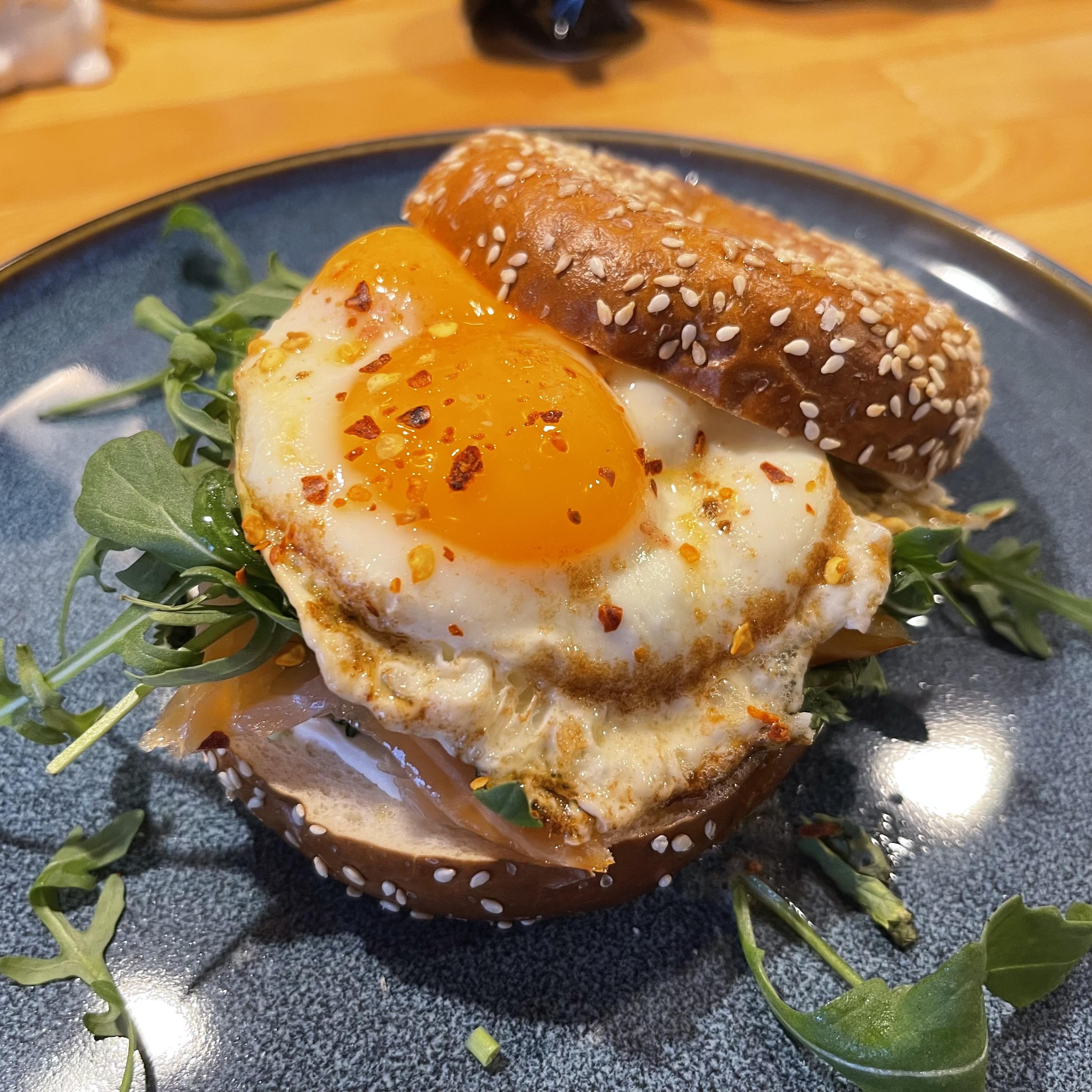 Na ciemnym talerzu leży przekrojony bajgiel z sezamem. W środku znajduje się wędzony łosoś i rukola, na wierzchu jajko sadzone z płynnym żółtkiem posypane płatkami chili. Talerz stoi na drewnianym blacie.