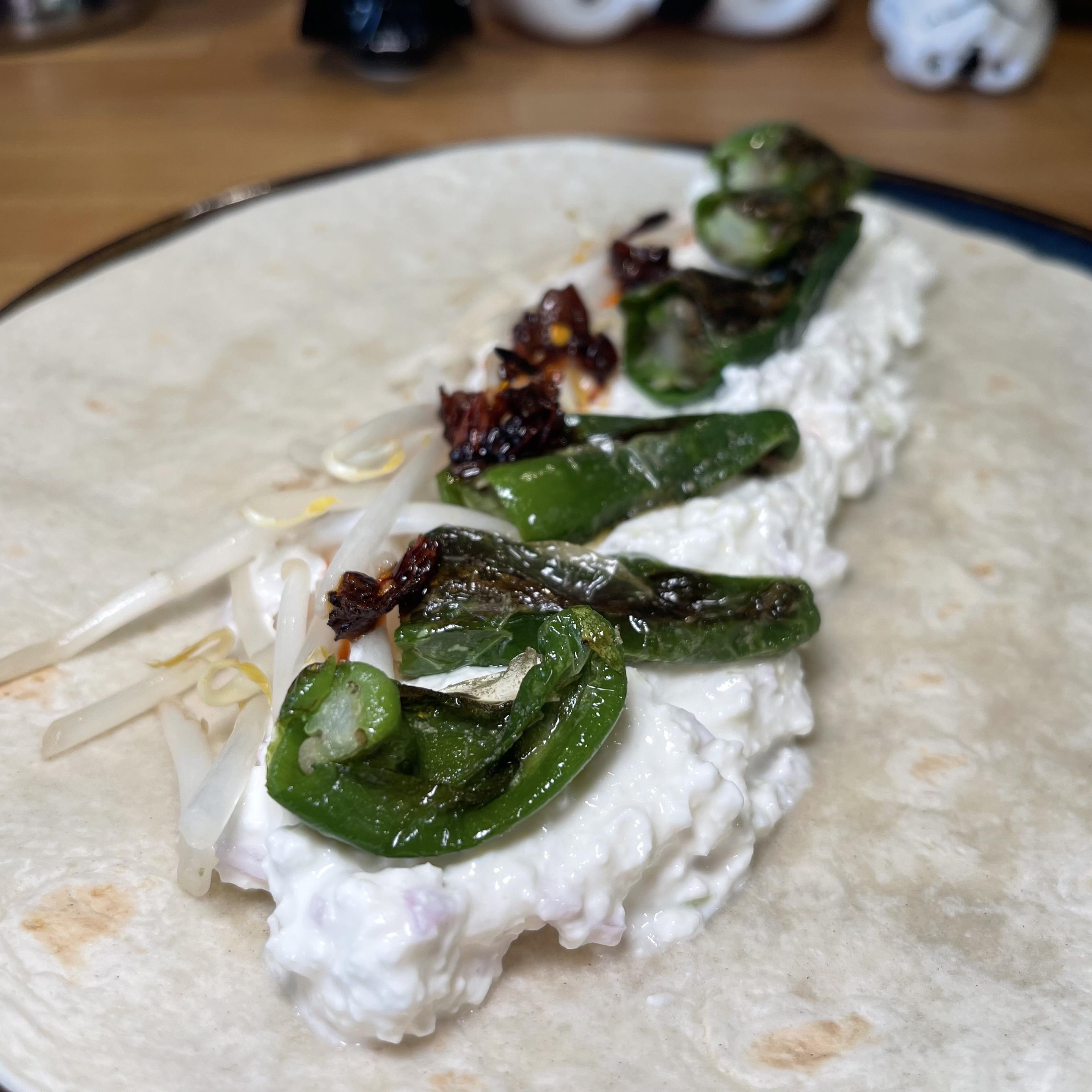 Na talerzu leży pszenna tortilla z grubą warstwą wytrawnego twarożku. Na wierzchu ułożone są podsmażone papryczki padrón, kiełki fasoli mung oraz kawałki chrupiącego chilli w oleju. Danie jest sfotografowane z bardzo bliska na drewnianym blacie kuchennym.