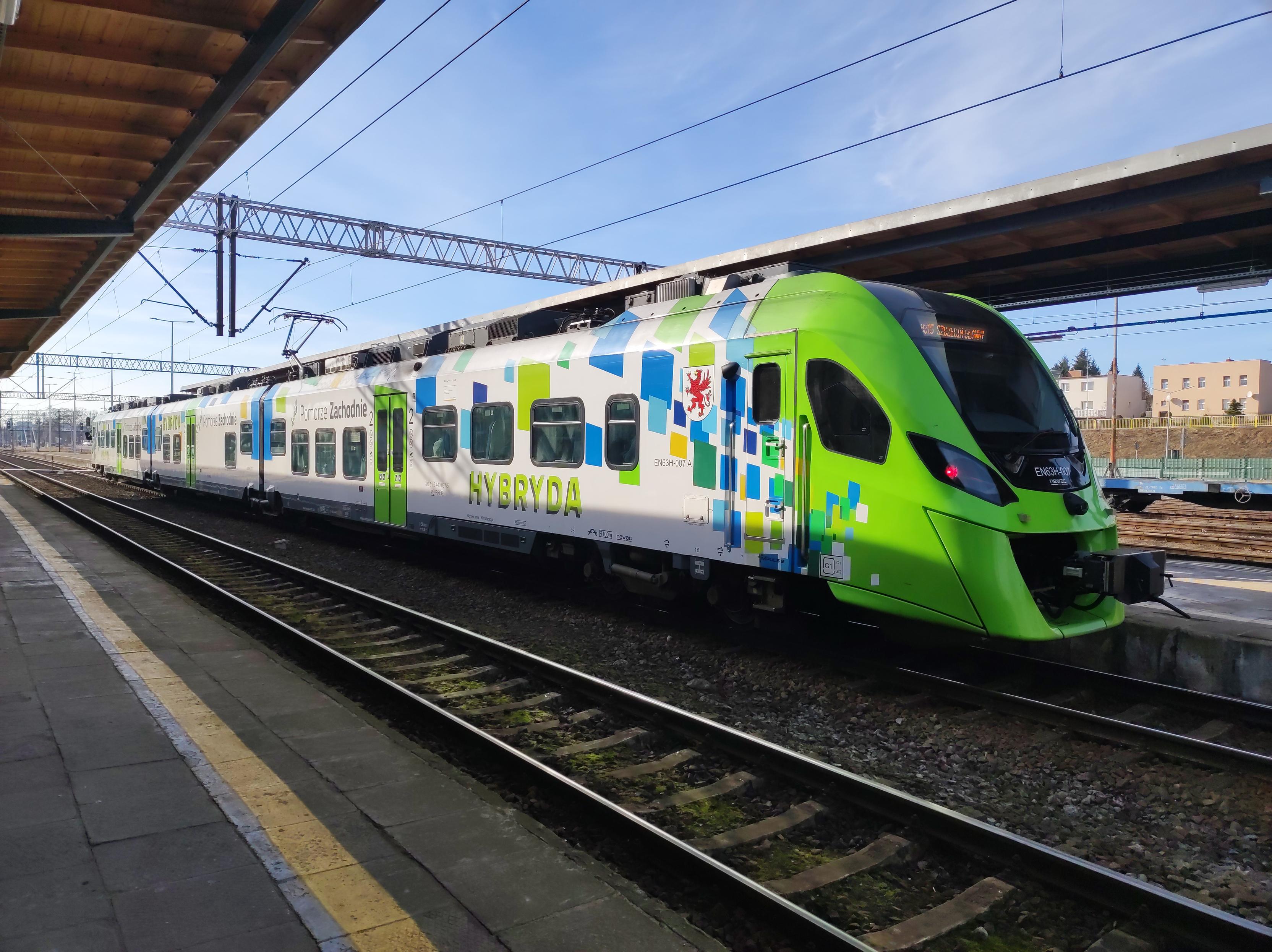 Biało-zielony EZT Newag Impuls stoi na peronie. Z boku ma zielony napis HYBRYDA i herb województwa zachodniopomorskiego - czerwonego gryfa.