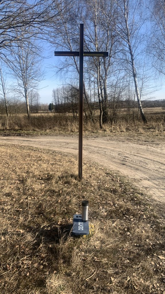 Metalowy krzyż na rozstaju piaszczystej drogi. Na betonowym słupku "poddupnik" Dexter, na nim duży kubek termiczny z kawą. Trawa jeszcze nie zielona. Pod lasem, w tle drzewa.