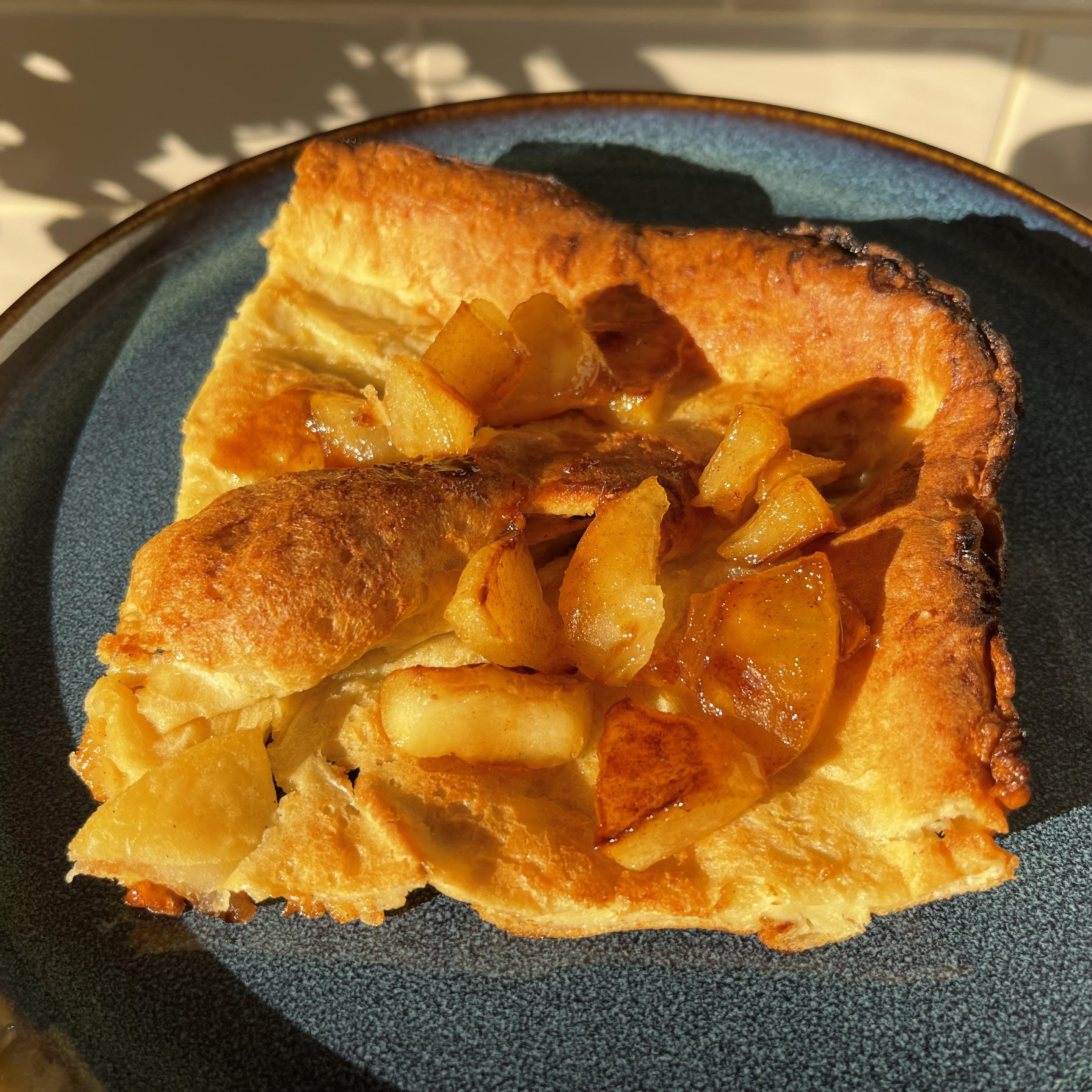 Na niebieskim talerzu leży kawałek puszystego pieczonego naleśnika typu Dutch baby z rumianymi, chrupiącymi brzegami. Na środku znajdują się kawałki jabłek karmelizowanych z cukrem i przyprawami. Całość oświetla ciepłe poranne słońce.