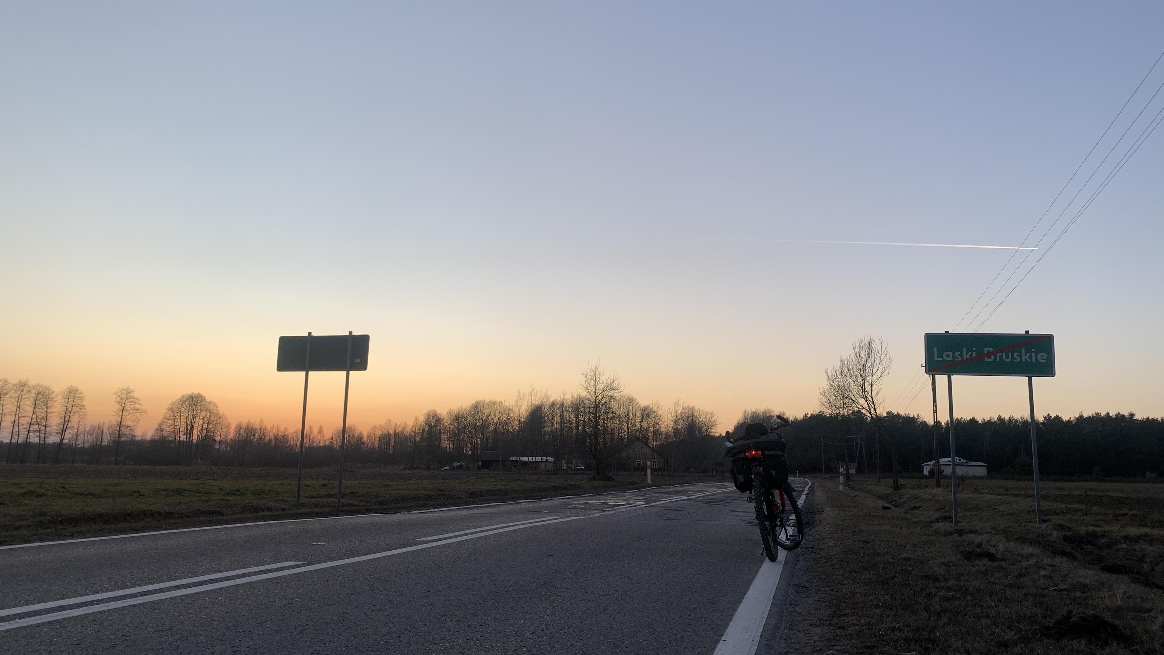 Asfaltowa droga na zachód, drogowskaz "opuszczasz Laski Bruskie", zachód słońca i las. Rower na poboczu
