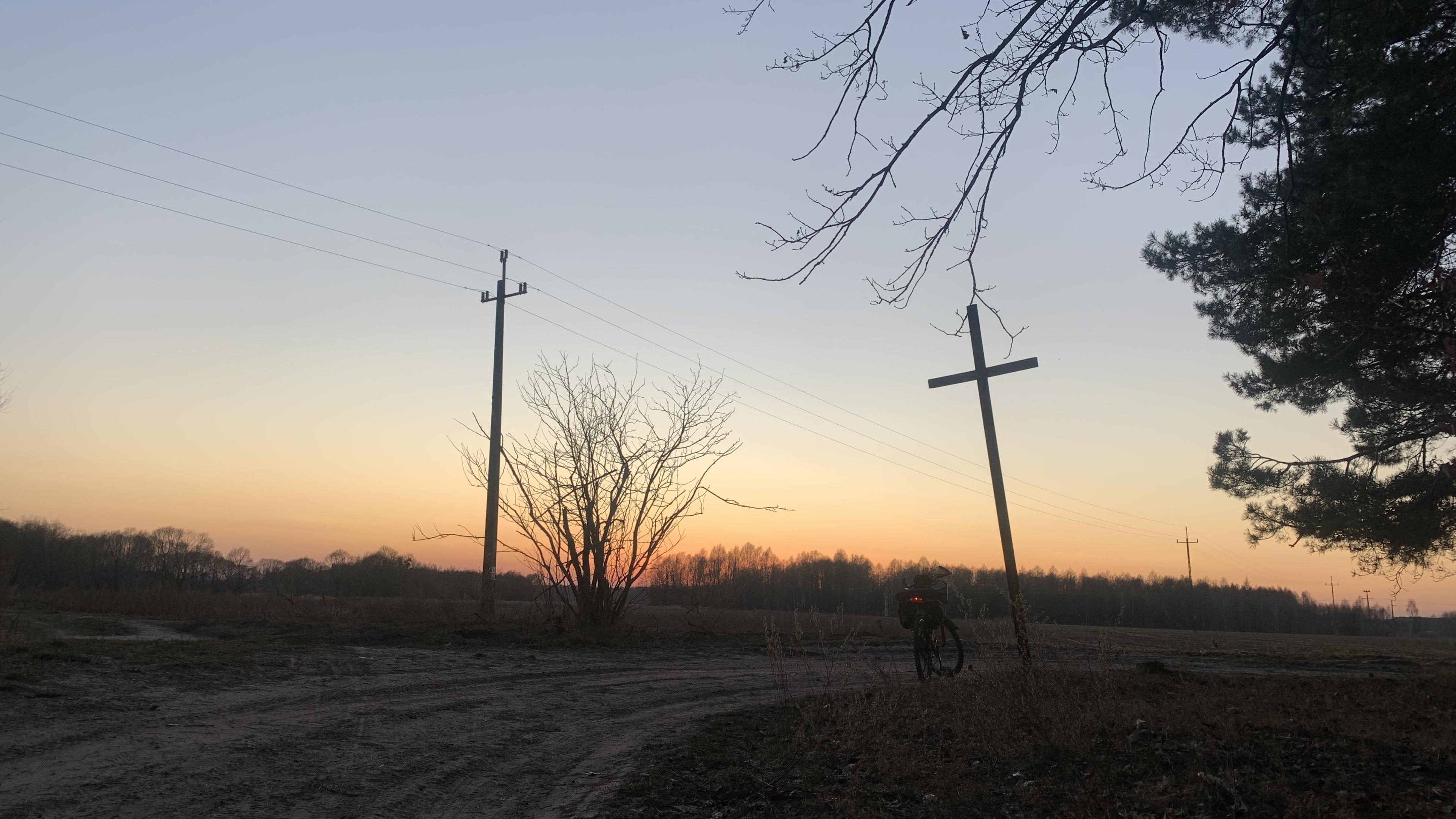 Rozstaje piaszczystych dróg, metalowy pochylony krzyż, obok słup energetyczny wyglądający jak krzyż, w tle jeszcze jeden. Zachód słońca nad lasem w tle. Obok krzyża rower