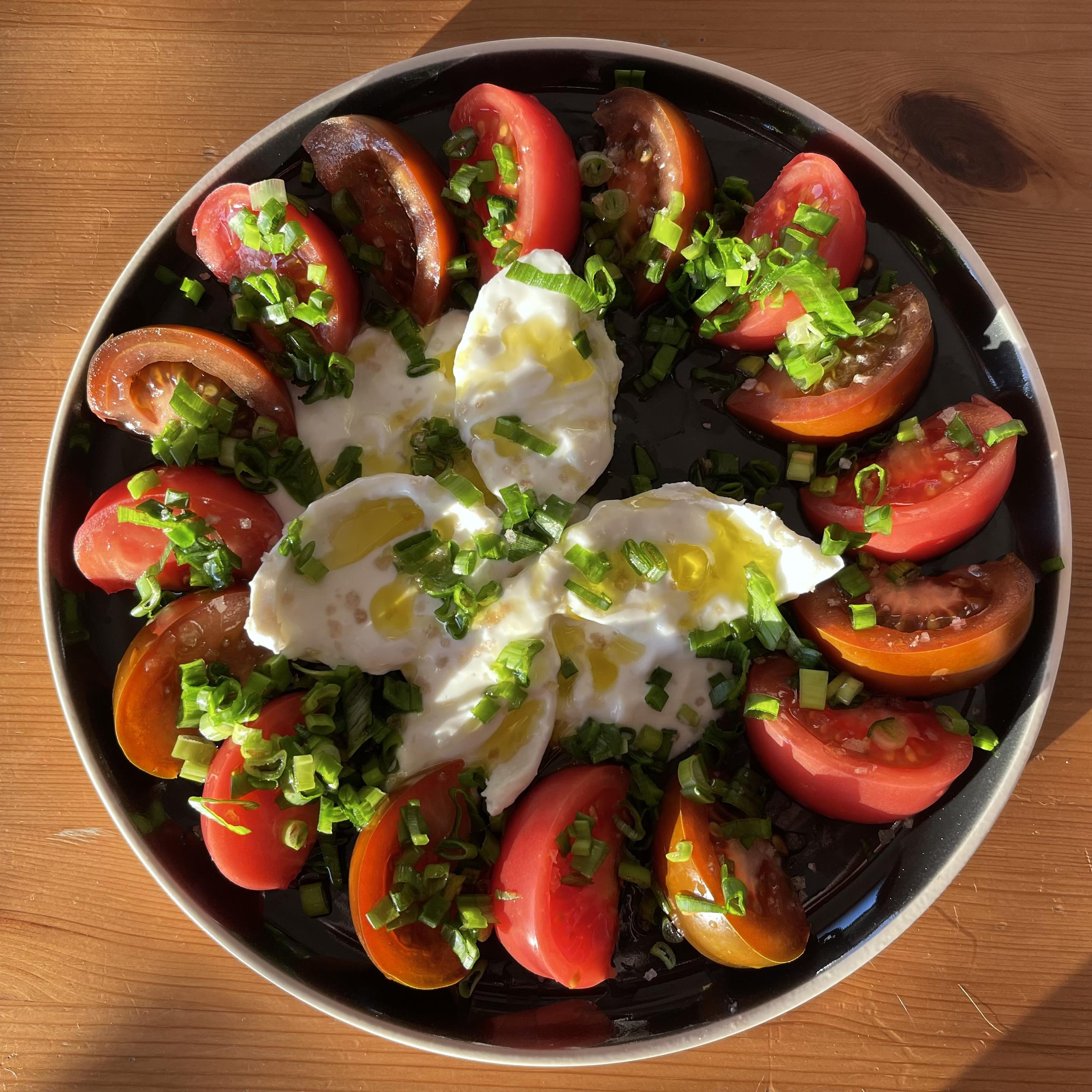 Na czarnym talerzu ułożone są ćwiartki pomidorów malinowych w różnych odcieniach czerwieni i brązu, tworzące okrąg wokół środka talerza. W centrum znajduje się rozerwana burrata z kremowym wnętrzem. Całość posypana jest drobno krojonym szczypiorem i polana oliwą z oliwek. Talerz stoi na drewnianym stole o ciepłym kolorze.