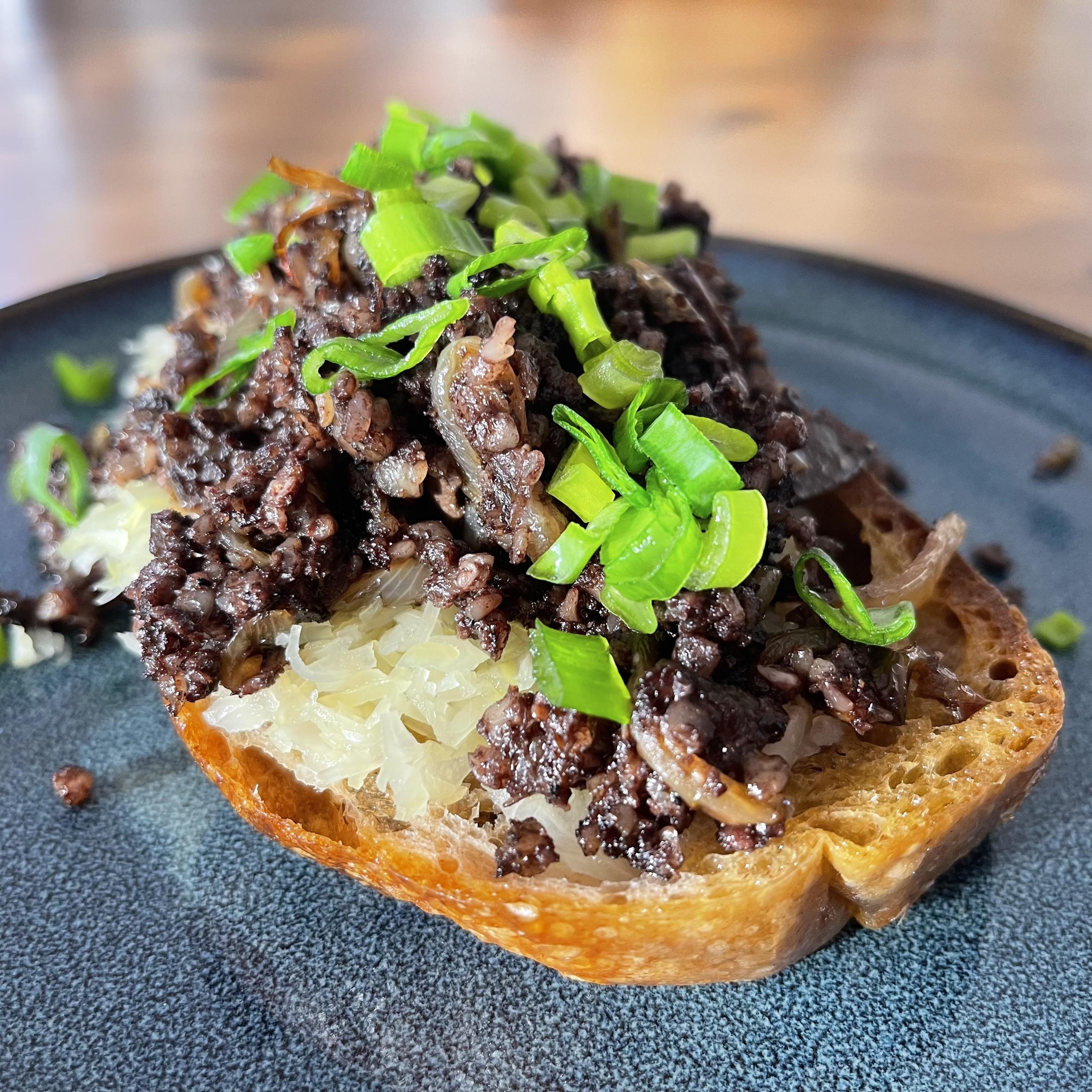 Na talerzu leży chrupiąca kromka pieczywa, na której znajduje się warstwa kiszonej kapusty, a na niej porcja podsmażonej kaszanki z kawałkami cebuli, posypana świeżym szczypiorkiem.