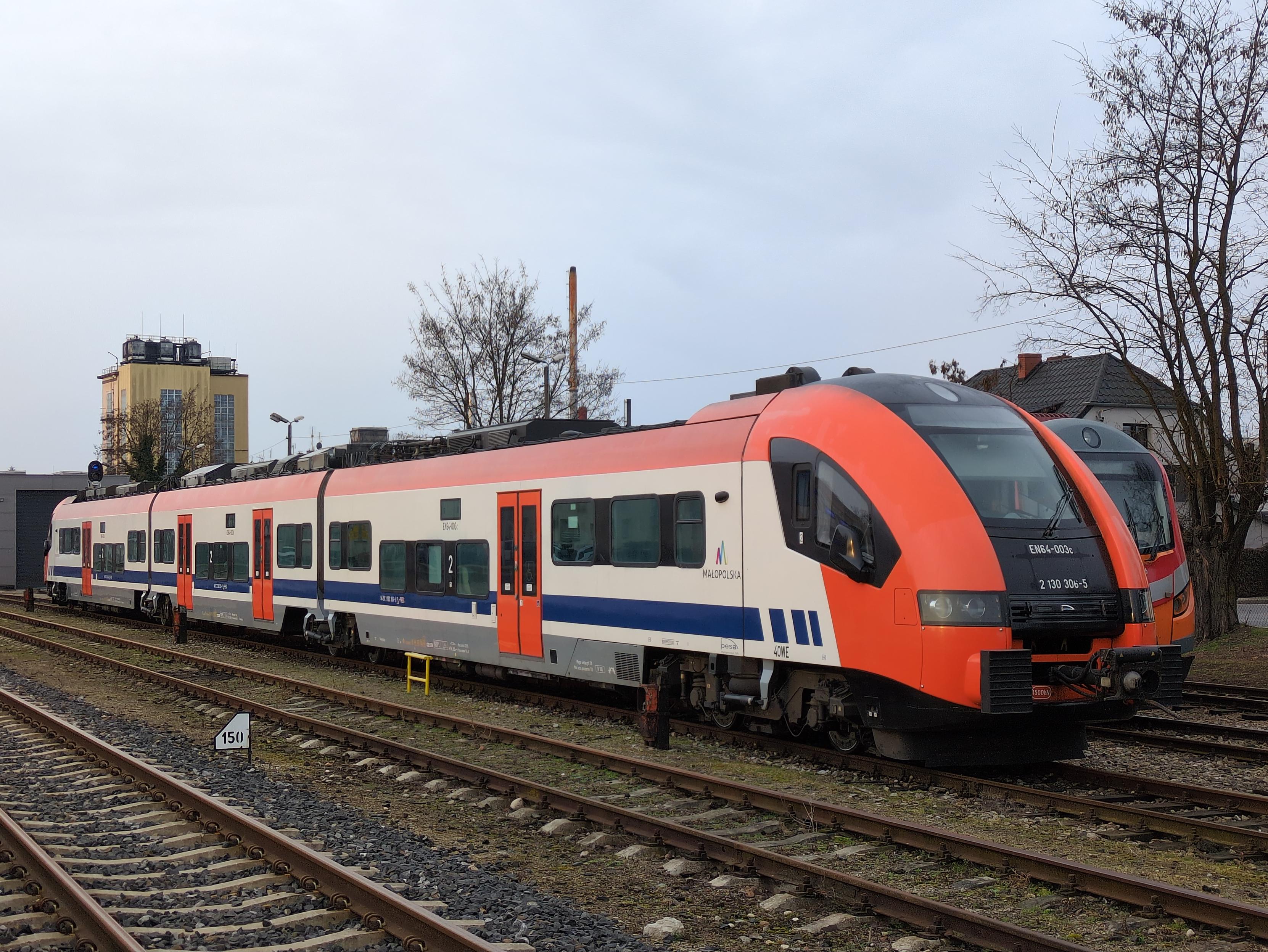 Trójczłonowy elektryczny zespół trakcyjny EN64-003 / Pesa 40WE (Acatus Plus), na bocznicy. Ma typowy, opływowy kształt z nieco wyższymi końcami. Malowany na biało, z czerwonym szczytem, przodem i drzwiami, i niebieskim pasem u spodu. Na boku napis "Małopolska", z kolorowym zygzakiem przypominającym literę M bądź góry. Za nim nieśmiało stoi pomarańczowo-czerwony kibel.