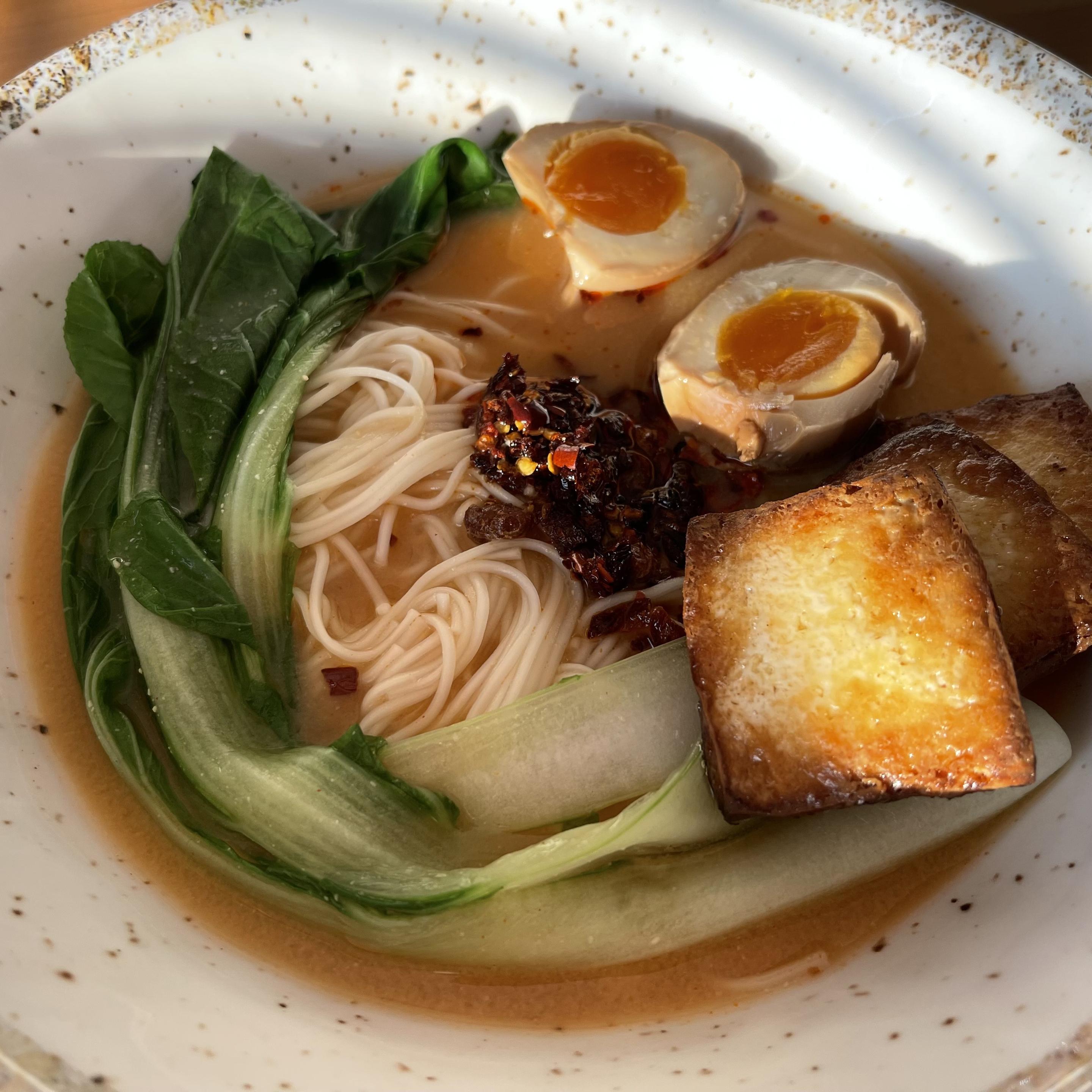 W głębokim jasnym talerzu podano miso ramen z jasnym bulionem, cienkim makaronem, dwoma połówkami marynowanego jajka z płynnym żółtkiem, liśćmi pak choi, kawałkami złocistego smażonego tofu i porcją chrupiącego chilli lao gan ma na środku