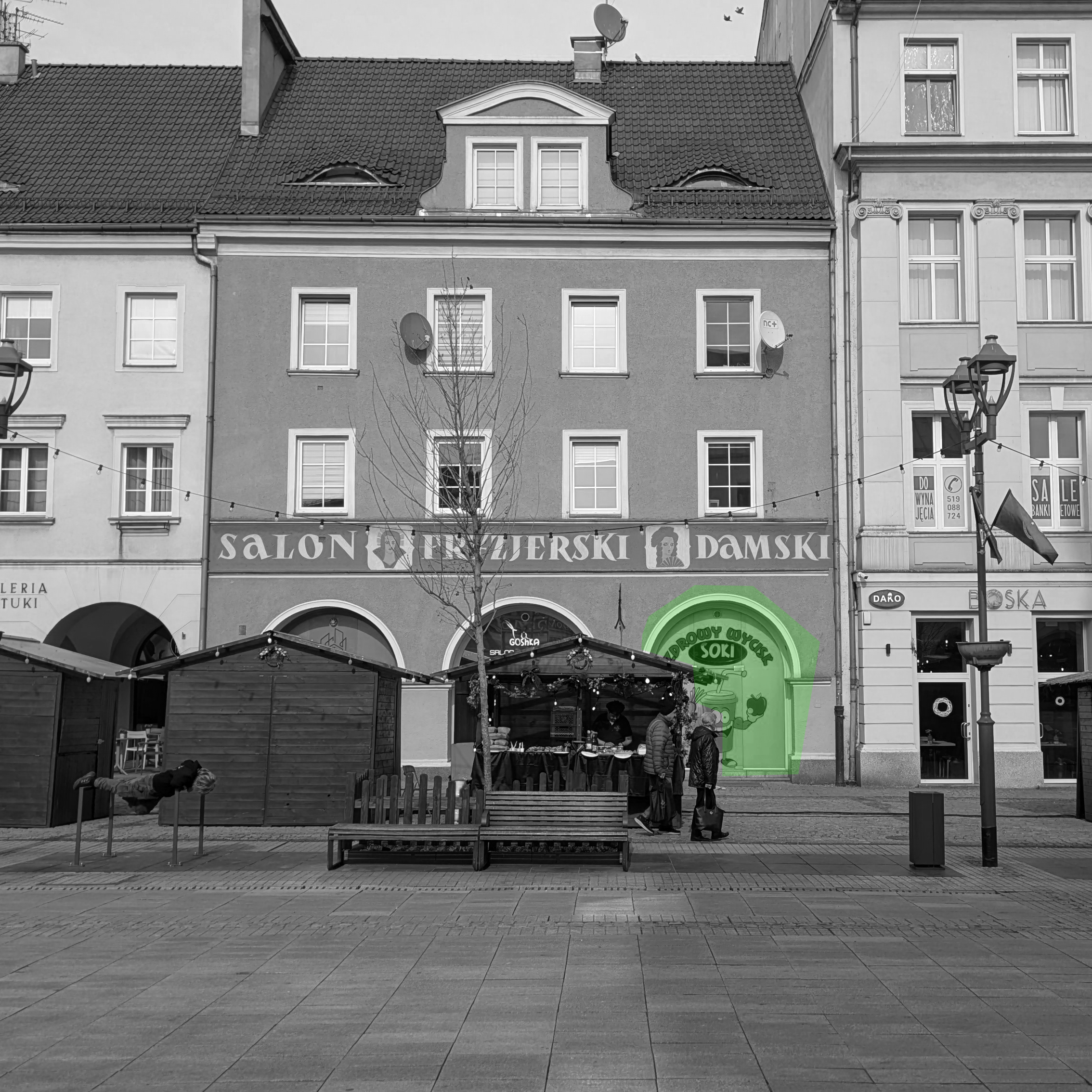 Zdjęcie budki na jarmarku wielkanocnym na tle pięknej kamienicy ze zdobieniami. W podcieniach kamienicy przeszklona witryna lokalu "Sokarnia", jaskrawozielono oklejona z marchwką która szczerzy zęby - reklama w stylu wczesnych lat dwuysięcznych.