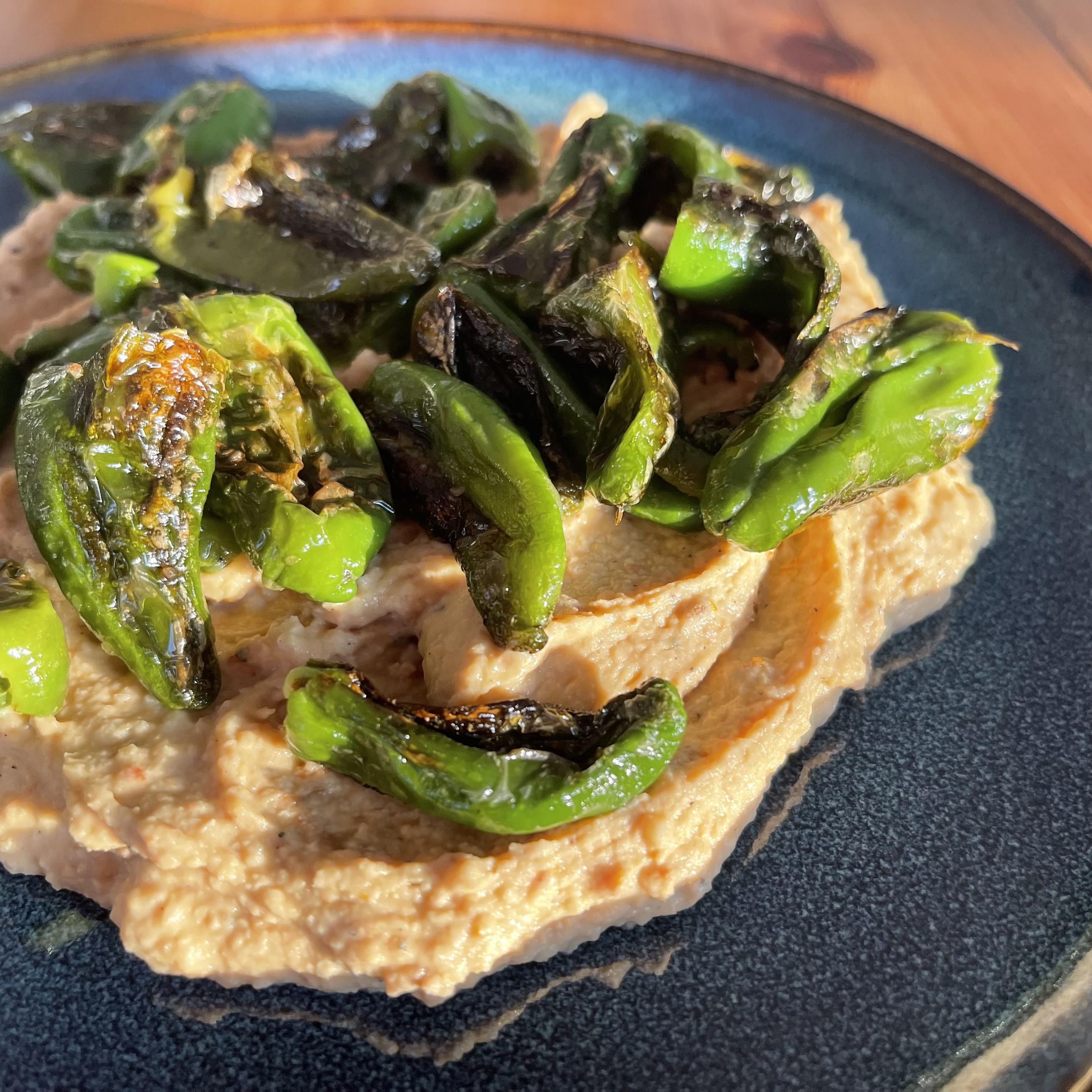 Na ciemnym talerzu znajduje się kremowy hummus z widocznymi kawałkami suszonych pomidorów, na którym ułożone są podsmażone papryczki padrone z lekko przypieczoną skórką.