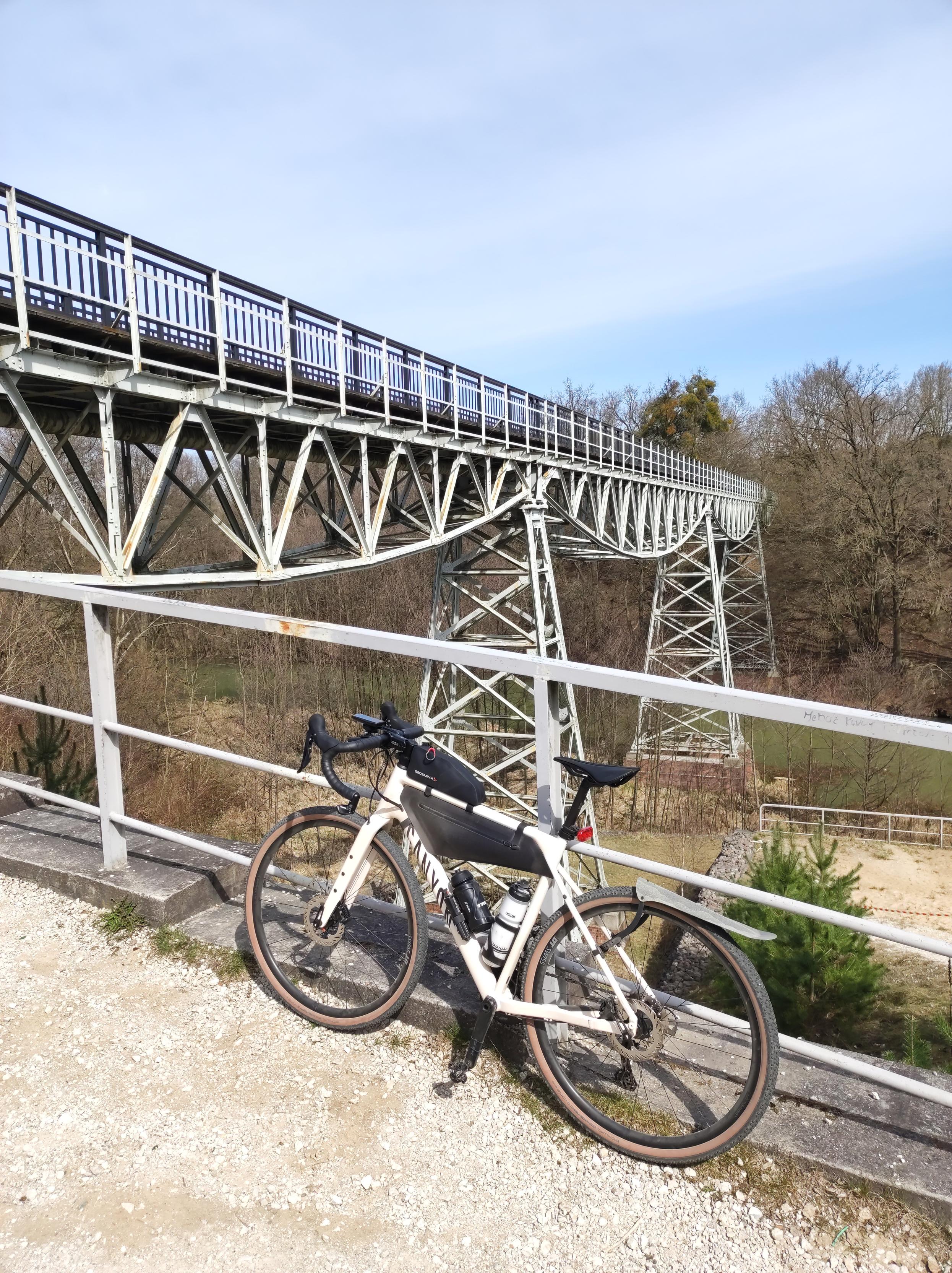 Kremowy rower szutrowy stoi oparty o metalową barierkę, a za nim wysoki most nad rzeką.