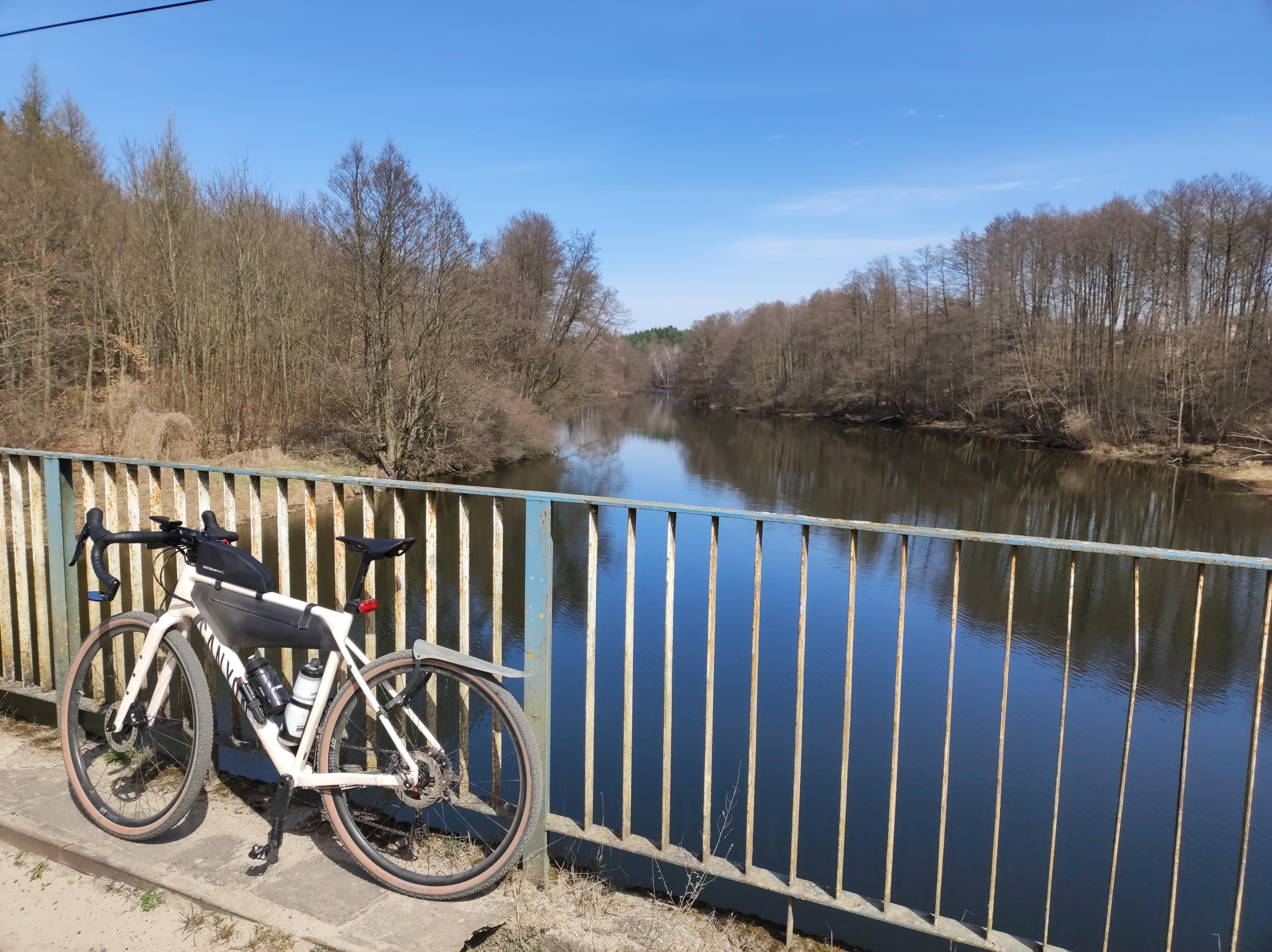 Mój rower oparty barierkę na moście nad Brdą, której brzegi porośnięte są drzewami i krzewami bez liści.