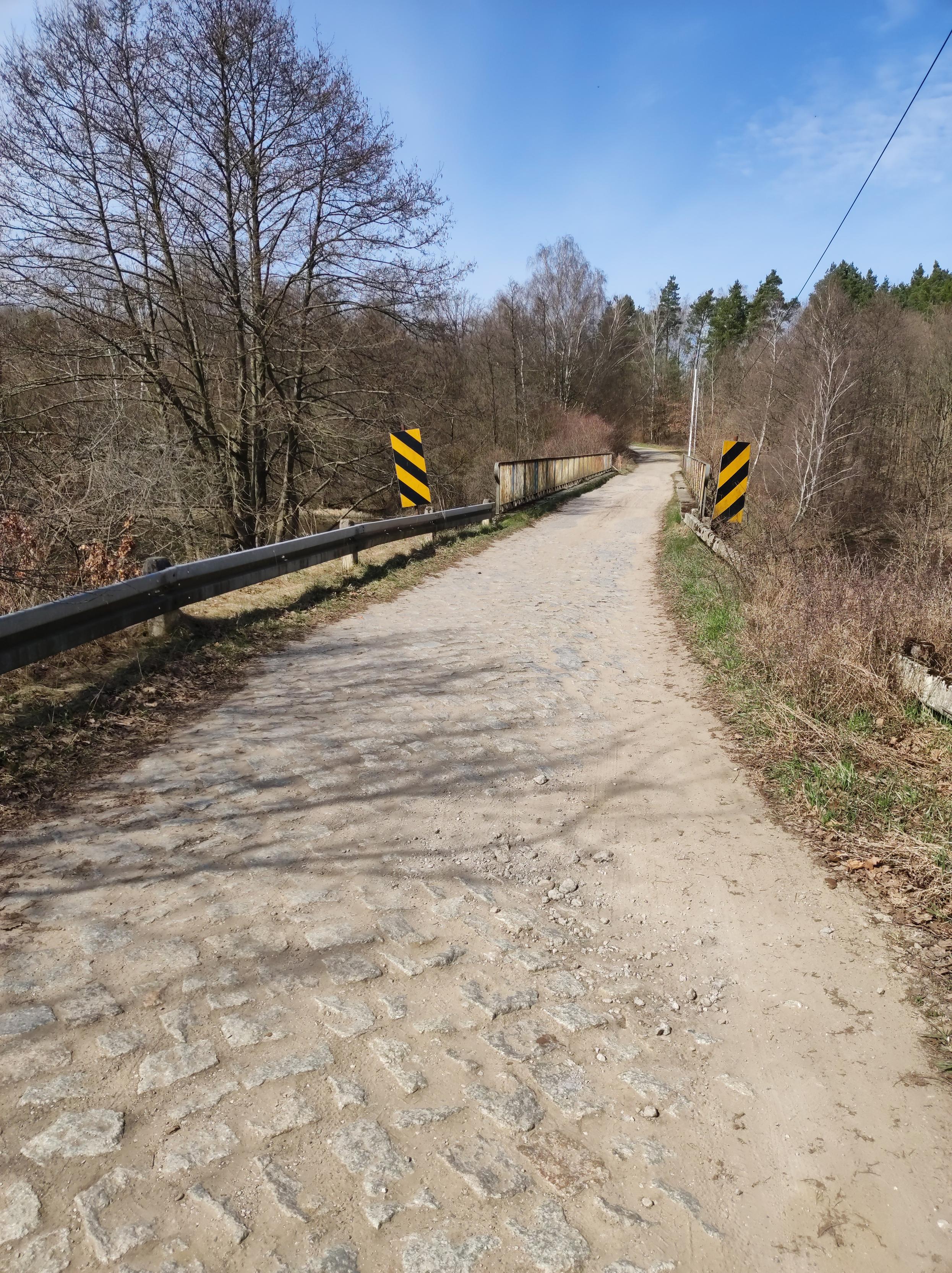 Wąska brukowana droga prowadzi do mostu na rzece z rdzewiejącymi barierkami.